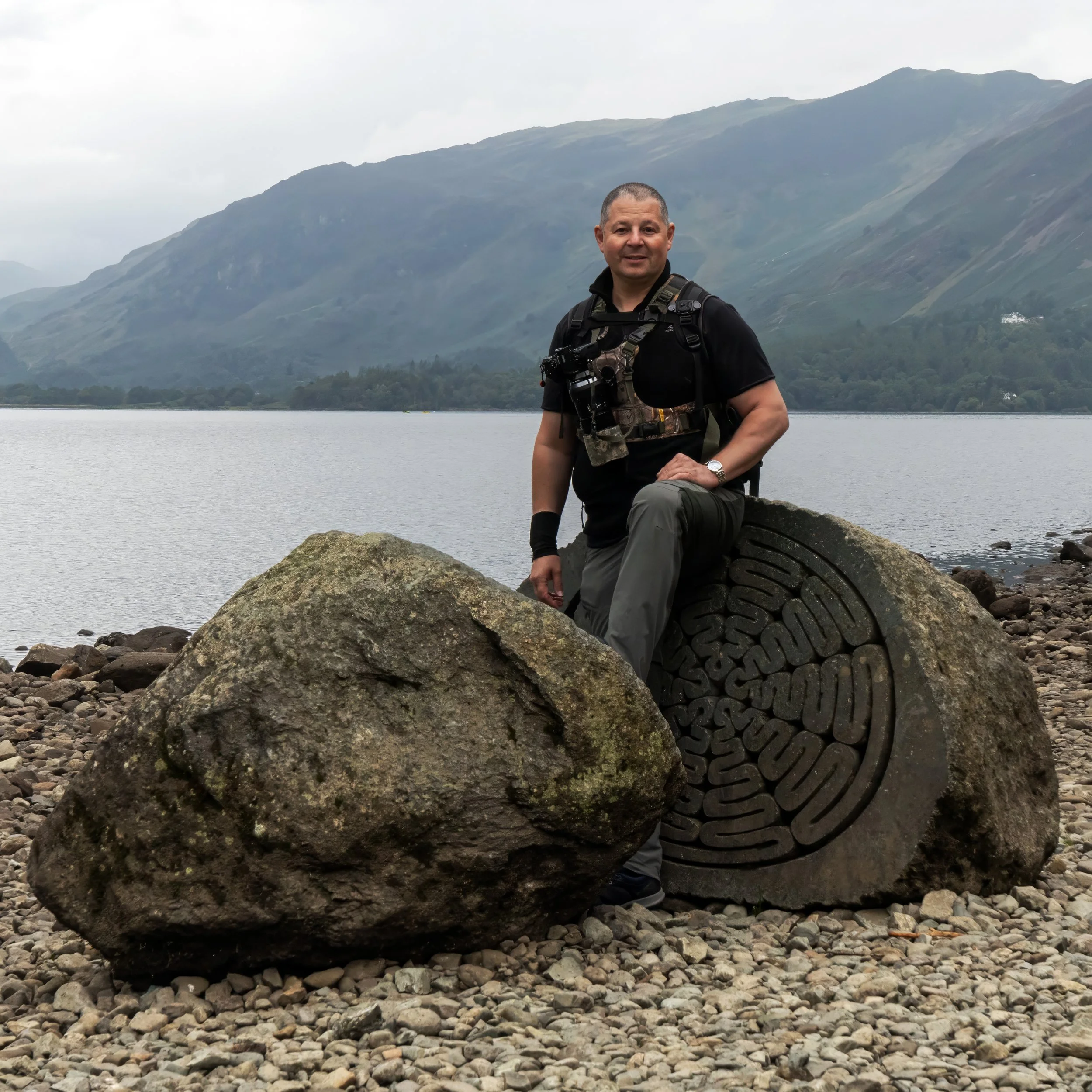 Man in outdoor gear sitting on a large rock by a lake with mountains in the background.
