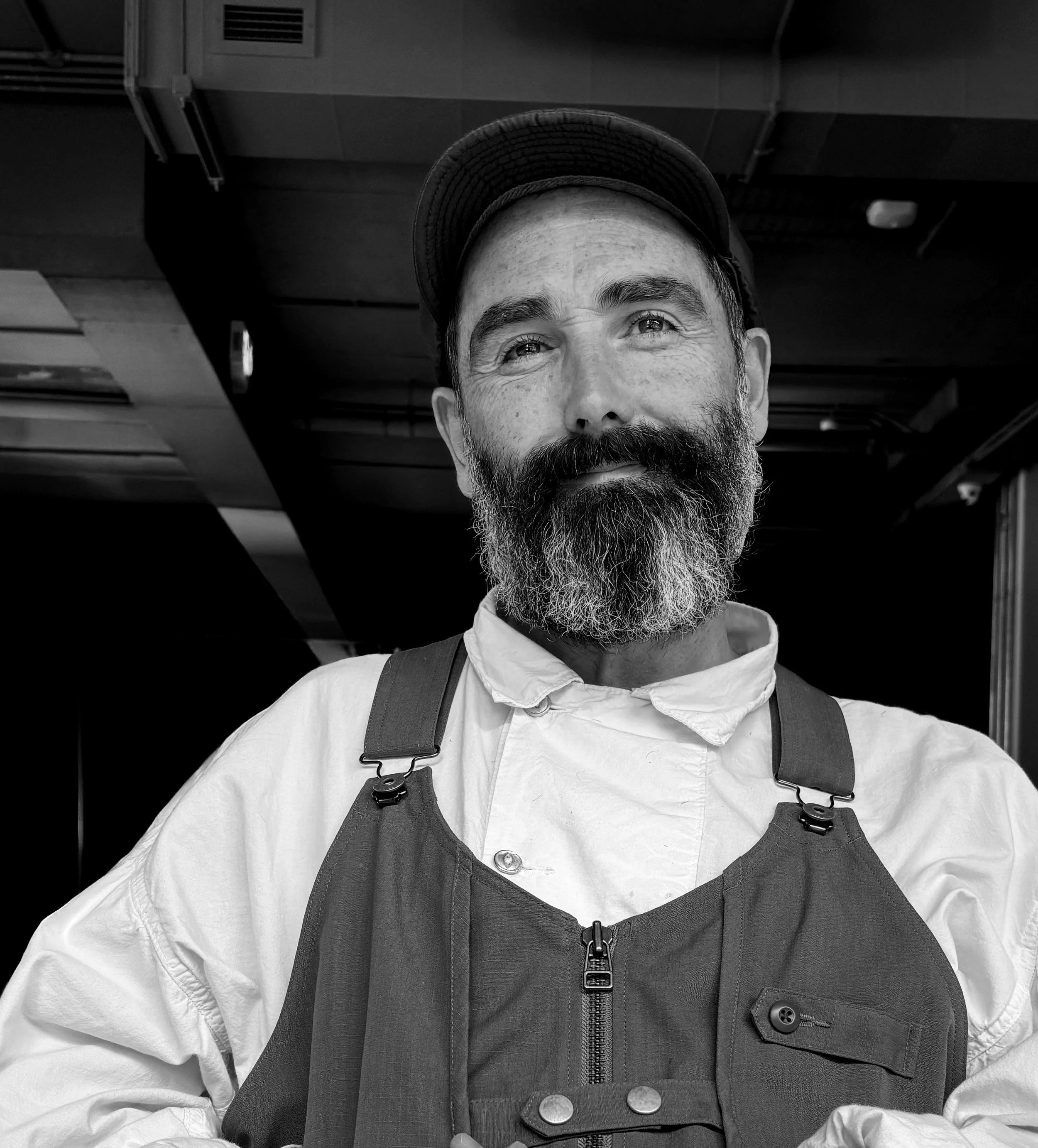 A man with a beard and mustache, wearing a dark cap, white chef jacket, and an apron, smiling in a professional kitchen setting.