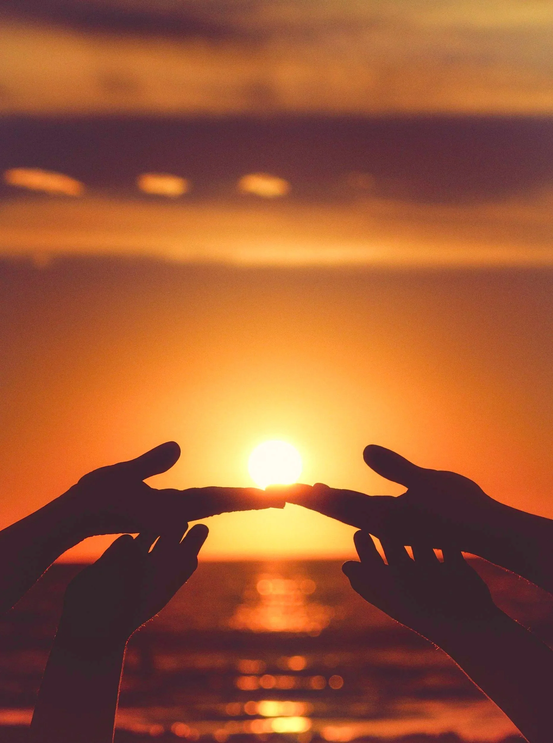 Silhouettes of two hands at sunset, with one hand touching the sun, over the ocean and colorful sky.