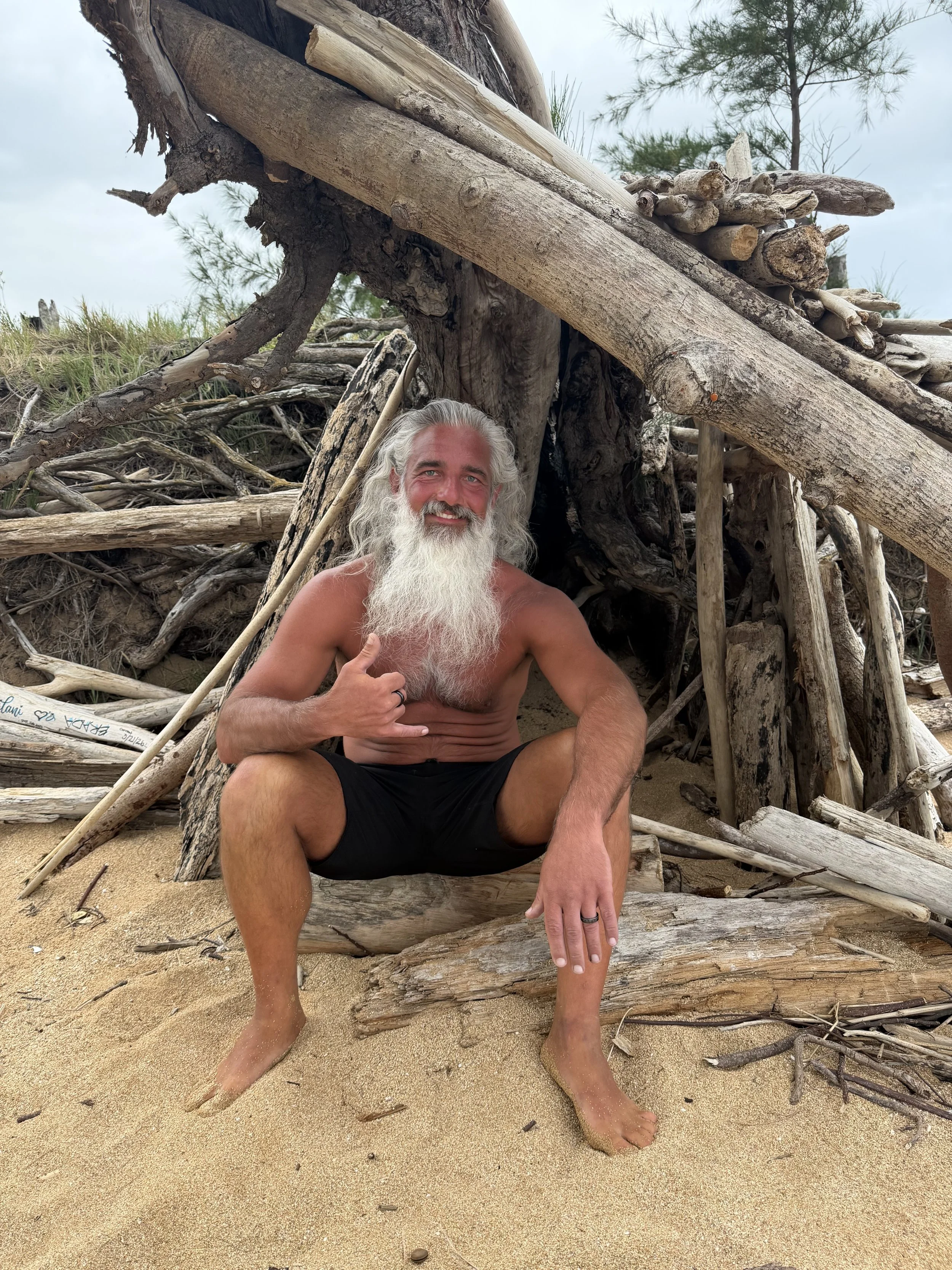 A shirtless man with long gray hair and a beard sitting on a piece of driftwood on a sandy beach, giving a thumbs-up and smiling, with a wooden structure made of driftwood and trees in the background.