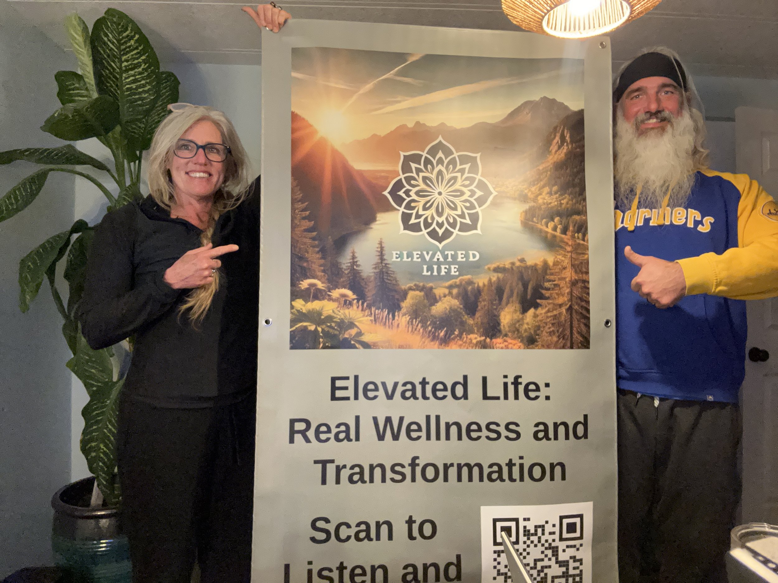 Two people, a woman and a man, holding a vertical sign with a nature landscape and a logo. The woman has blonde hair, glasses, and wears a black jacket. The man has a beard, long hair, a black beanie, and a blue and yellow jacket. The sign reads "ELEVATED LIFE" with a mountain and lake scene, and a QR code at the bottom. There is a large green plant on the left and a woven light fixture above.