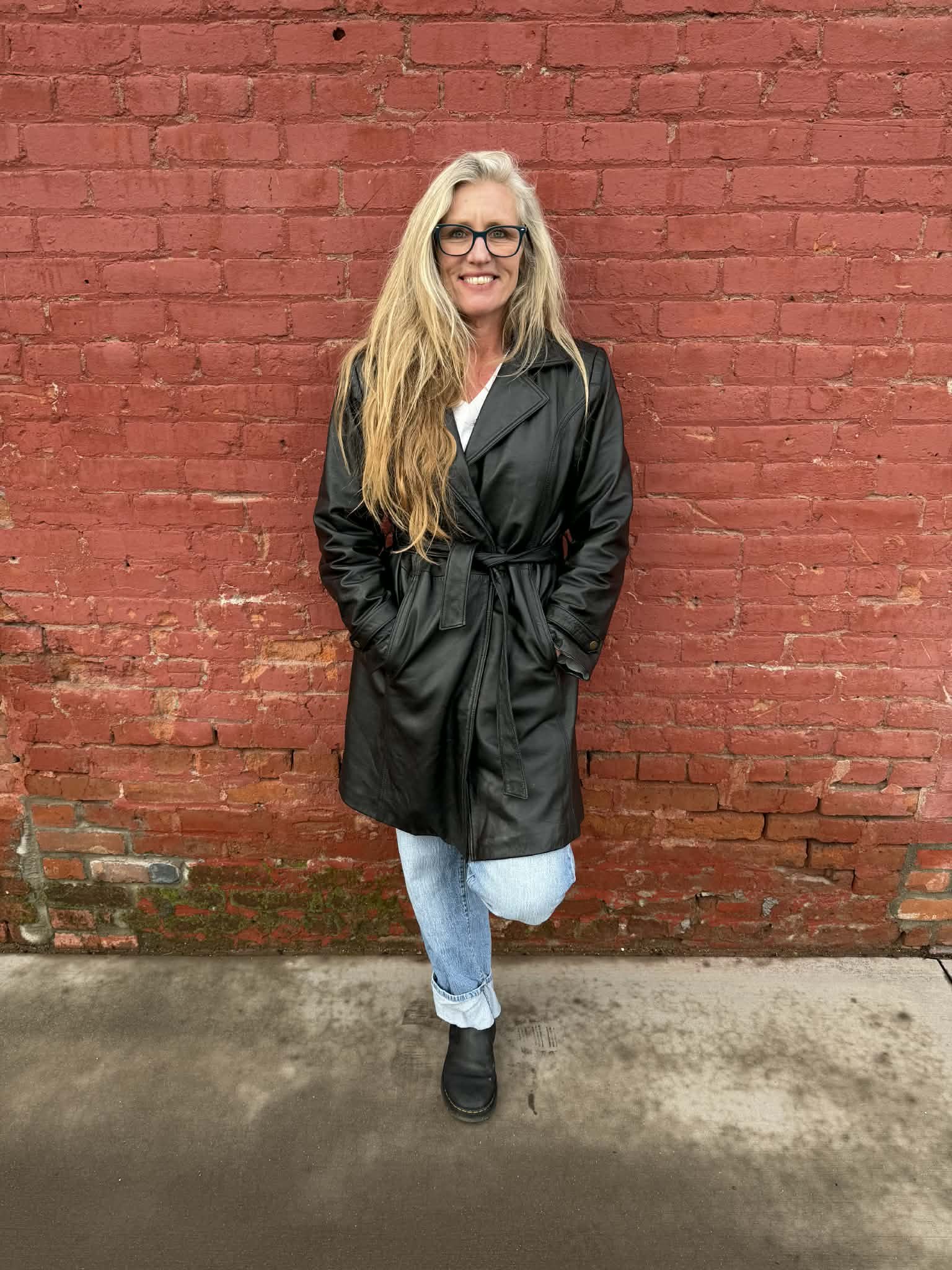 A woman with long blonde hair, glasses, wearing a black trench coat, light blue jeans, and black boots standing against a red brick wall.