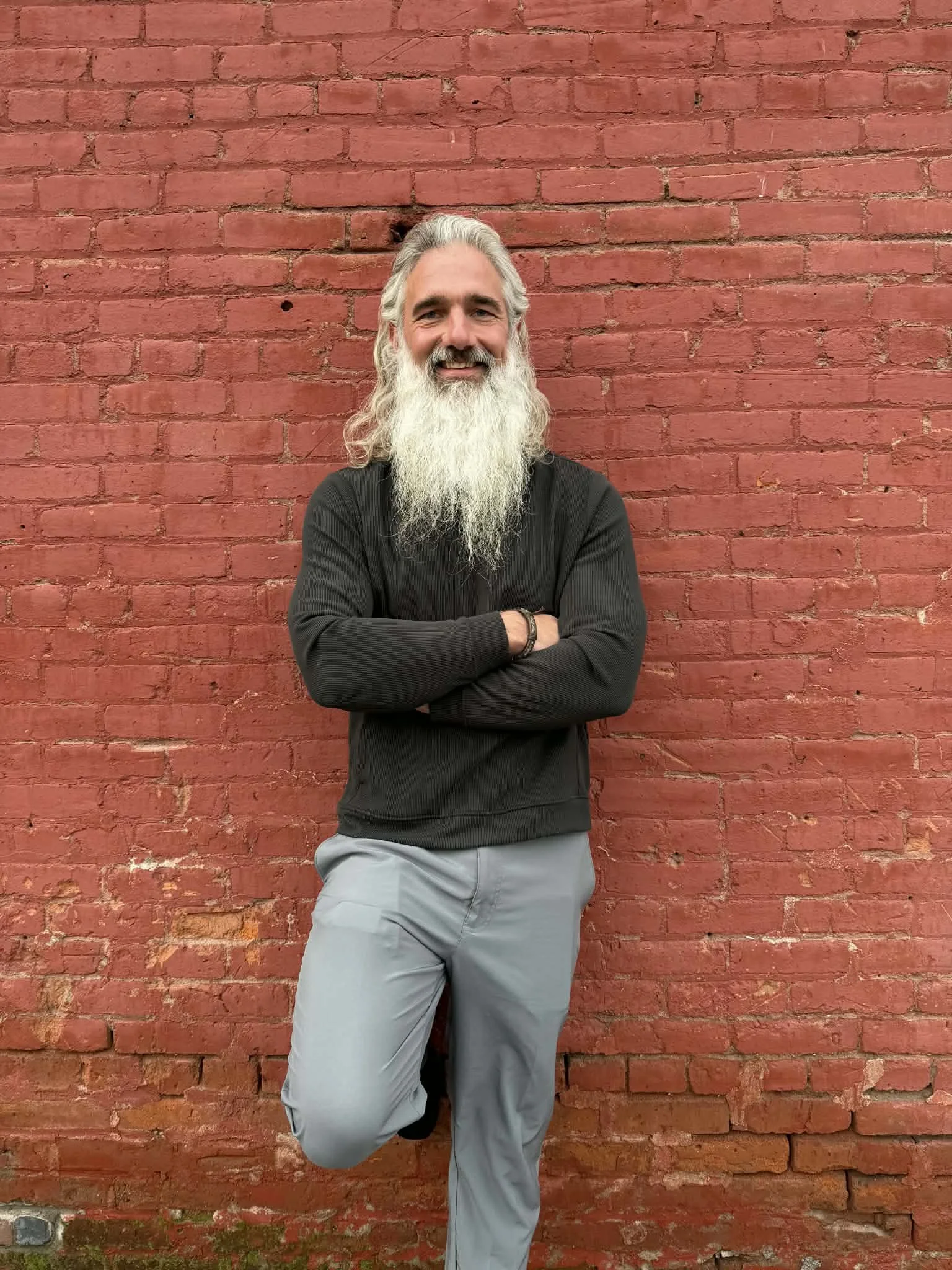 A man with long gray hair and a full gray beard, smiling, standing against a red brick wall with arms crossed and one leg bent, wearing a black long-sleeve shirt and light gray pants.