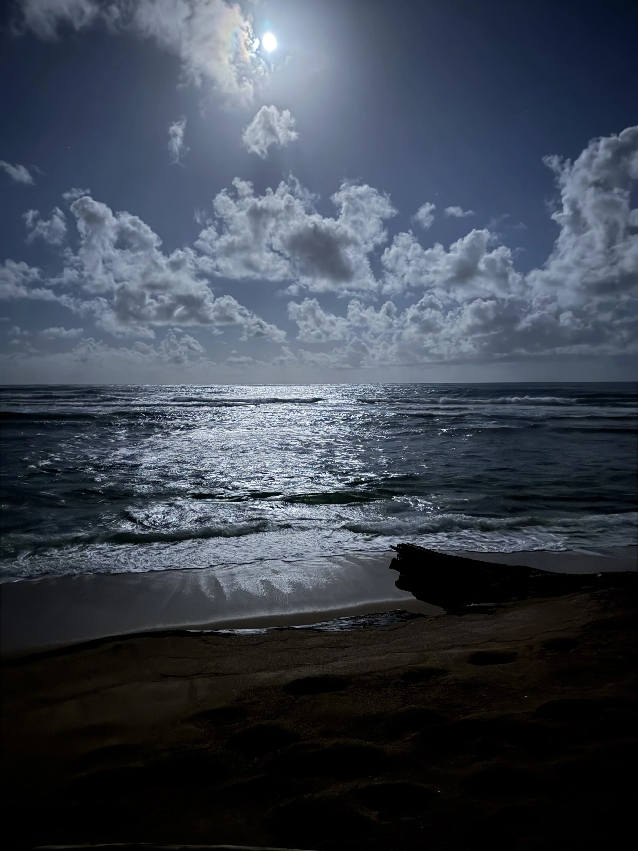 A nighttime scene at the beach with dark ocean waves, reflections of moonlight on water, partly cloudy sky, and a bright full moon partially covered by clouds.