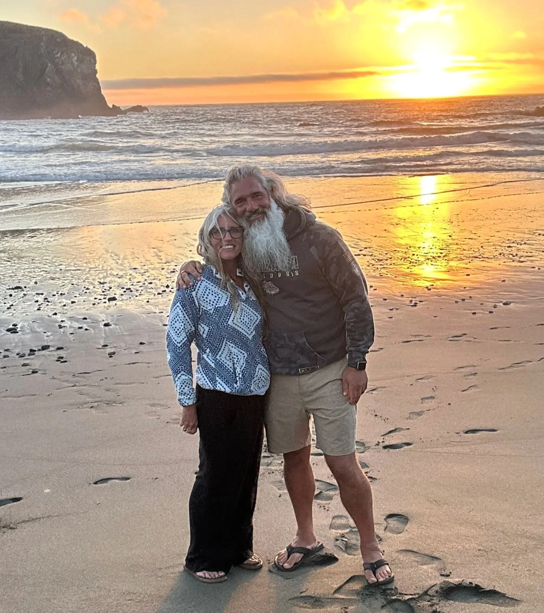A couple with gray hair and beards stands on a beach at sunset, smiling, with a cliff and the ocean in the background.