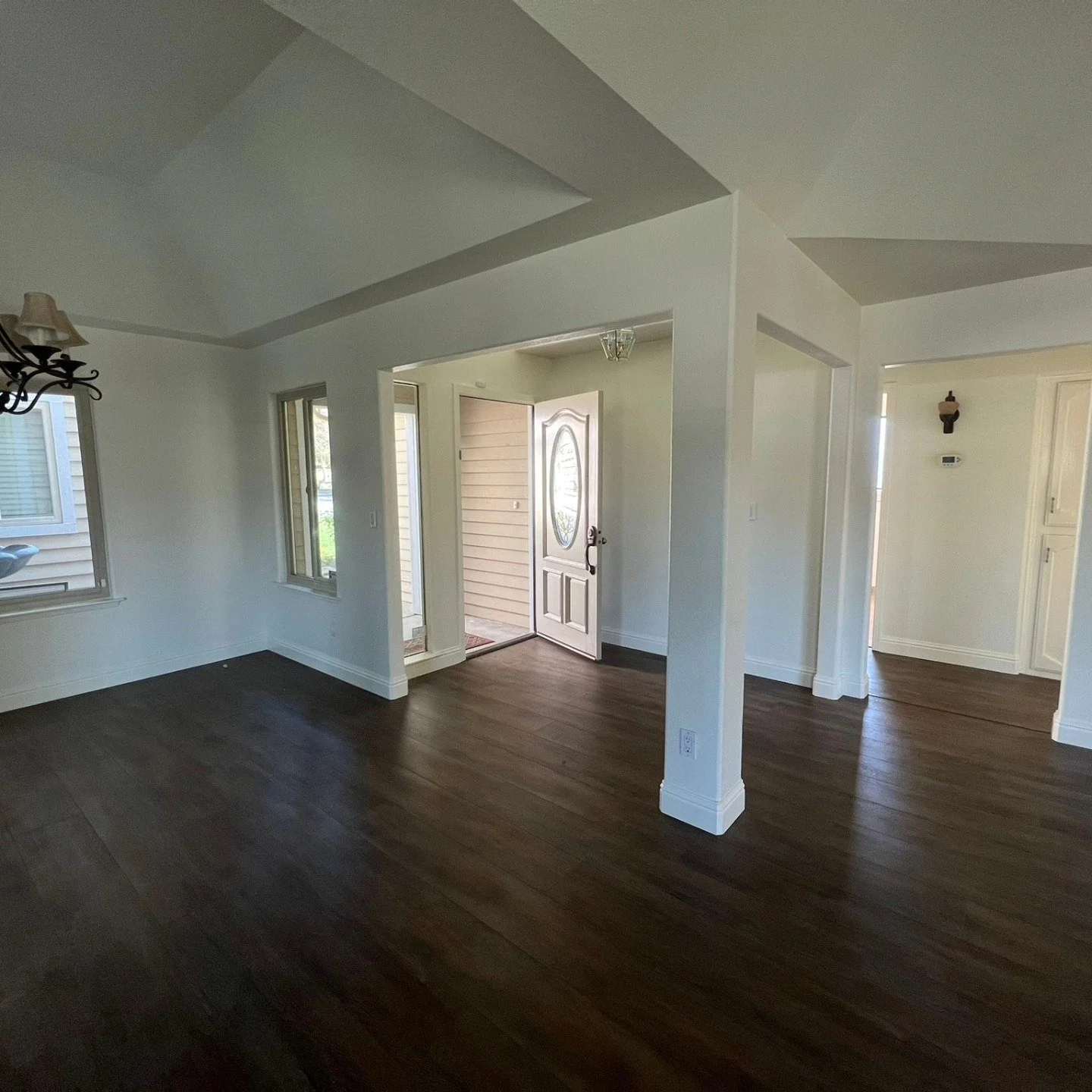 Empty living room with dark hardwood flooring, white walls, windows, and an open front door leading outside.
