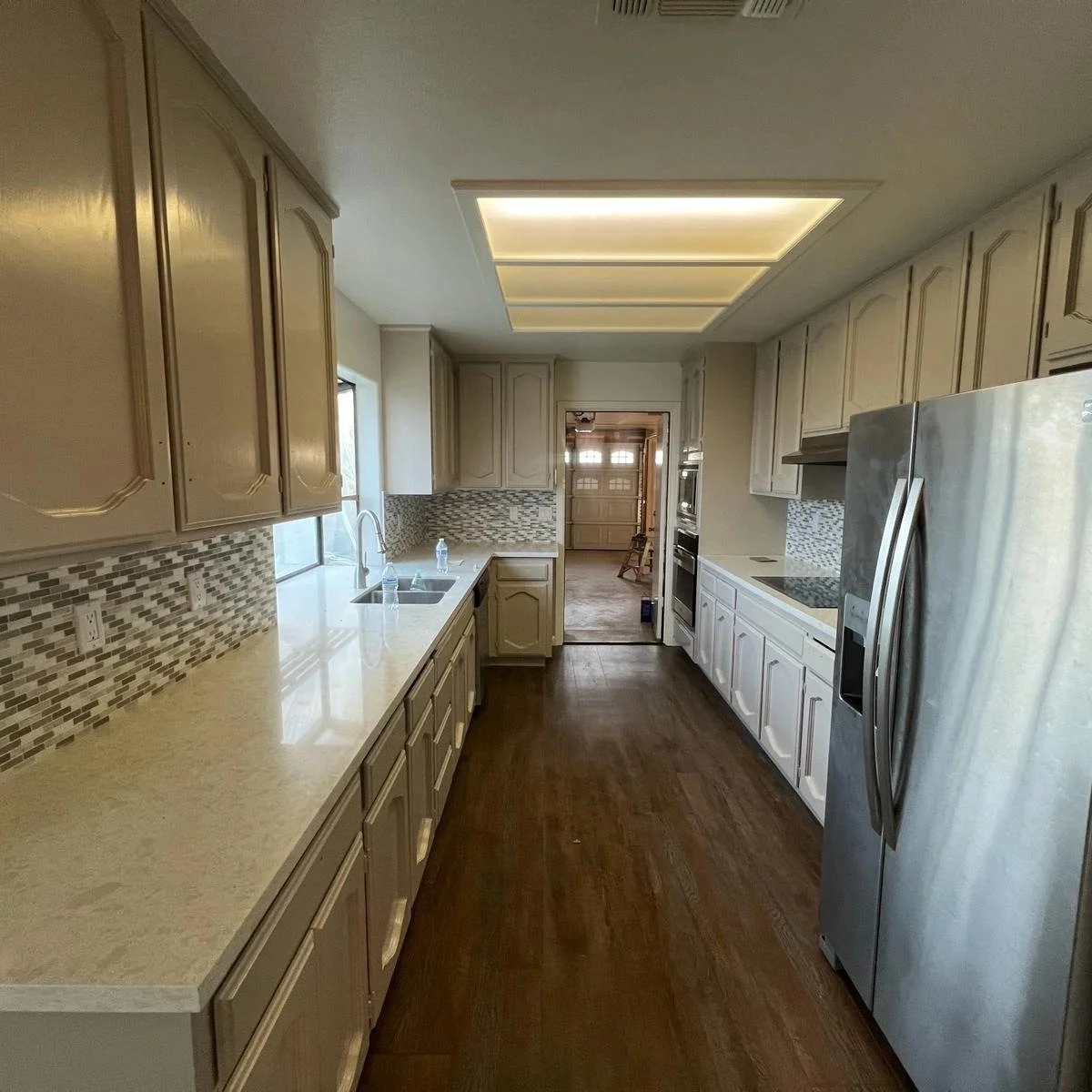 Kitchen with beige cabinets, a mosaic tile backsplash, a double sink under a small window, and stainless steel appliances including a refrigerator and oven. The ceiling has recessed lighting, and there is a doorway leading to another room with a gara