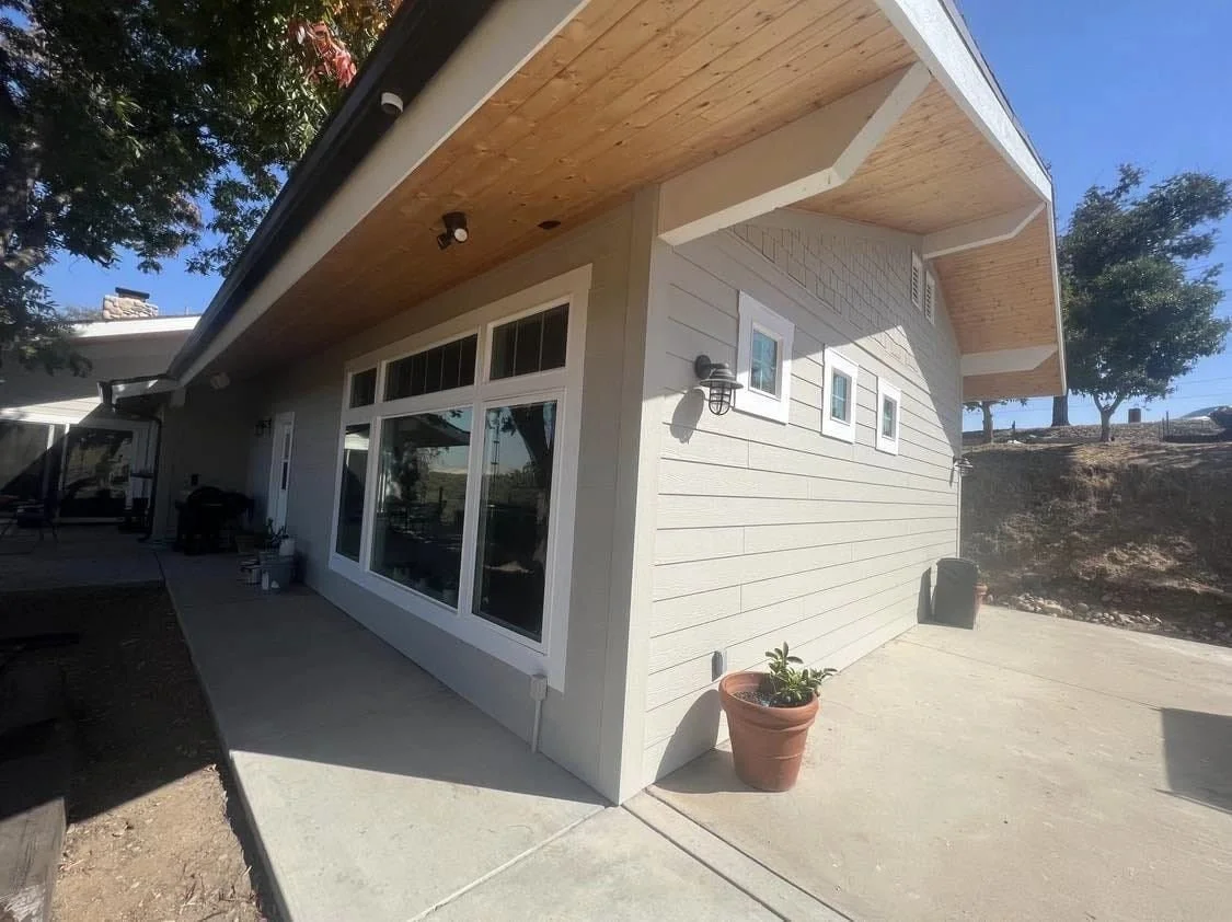 Exterior view of a modern house with beige siding, large windows, and a wooden overhang on a sunny day.