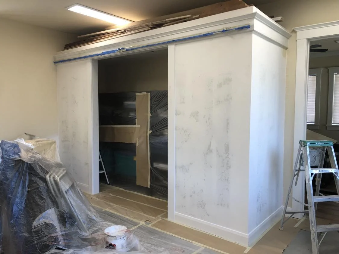 Room under construction or renovation with plastic and paper covering on the floor and furniture, a partially painted wall with painter's tape, and a step ladder