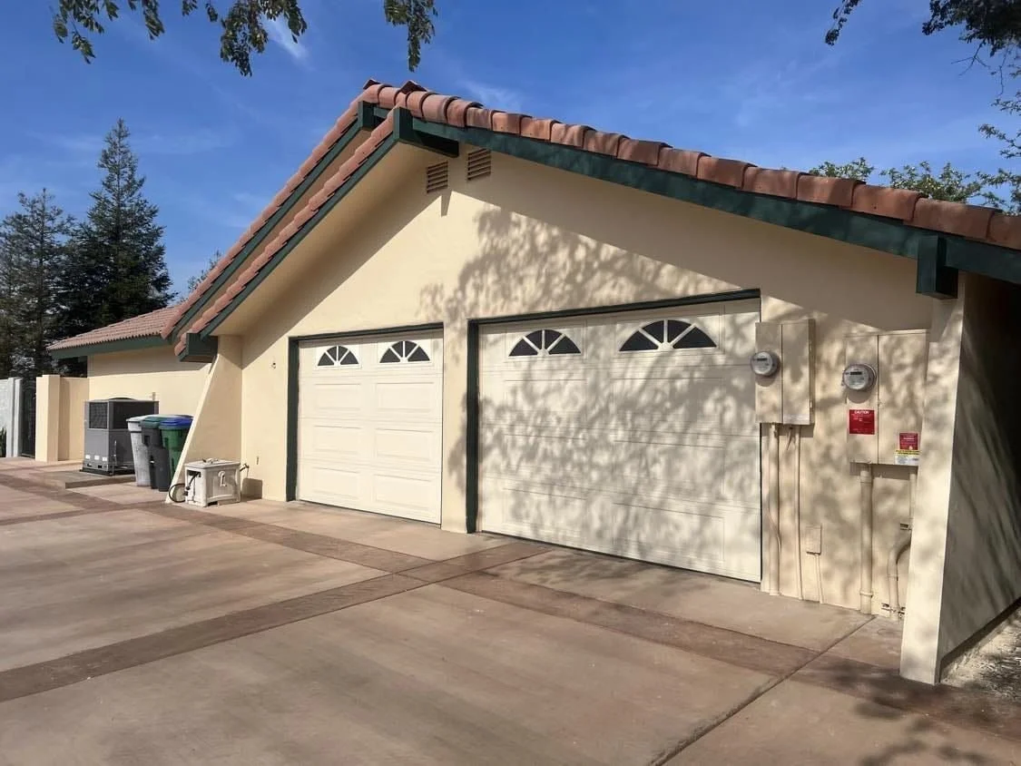 Two white garage doors with small windows in a beige building with a tiled roof, under a blue sky, with trees in the background.