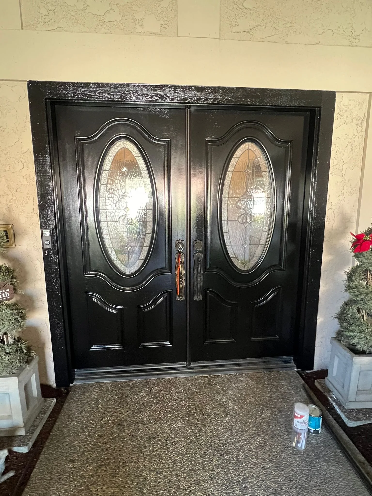 Black double front door with oval stained glass windows, decorated with holiday plants on each side, and a small container and paint supplies on the floor nearby.