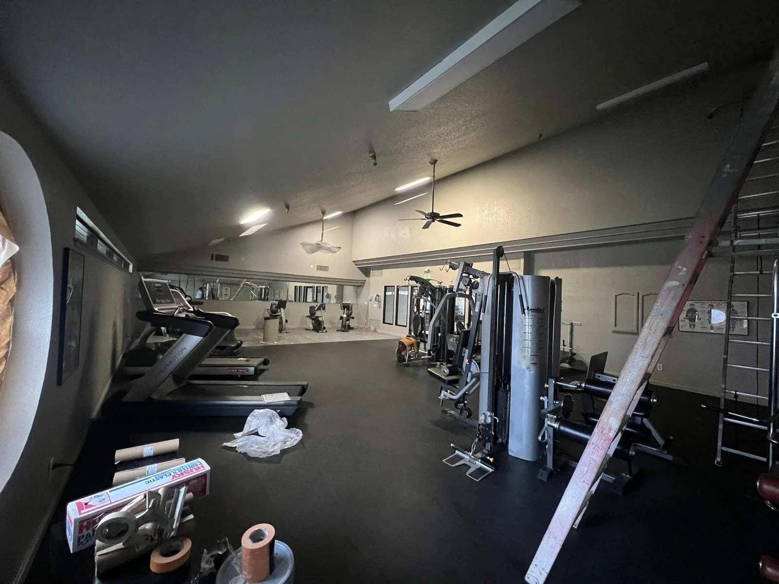Empty gym with treadmills, weight machines, and exercise bikes, with some construction or maintenance items and unfinished areas.