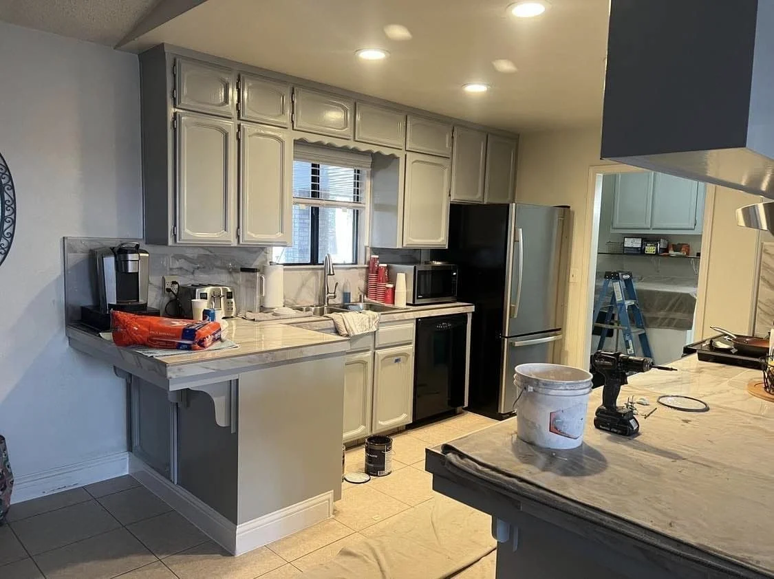 A kitchen with white cabinets, a window above the sink, and various appliances including a refrigerator, microwave, and coffee maker. There are items on the countertops, and a bucket and tools on a nearby island or counter, indicating ongoing kitchen
