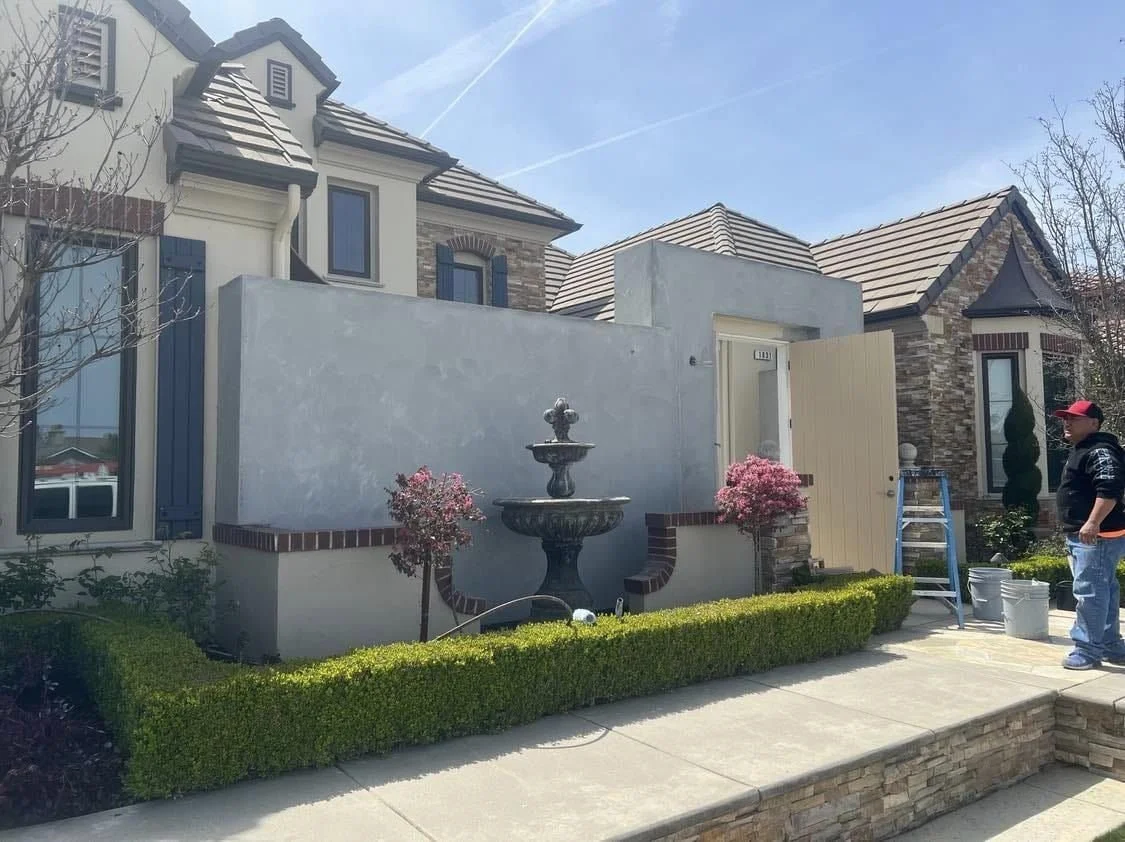 A man standing outside a modern house under construction, with a central decorative fountain, pink flowering bushes, and neatly trimmed green hedge, in a sunny residential neighborhood.