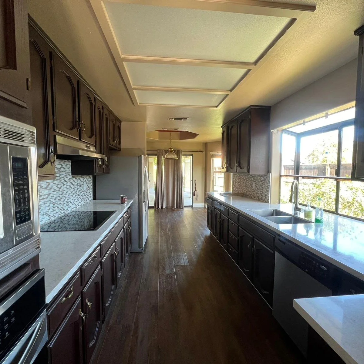 A kitchen with dark wooden cabinets, white countertops, a stainless steel microwave, and a refrigerator. There is a double sink near a large window with a view of trees and a backyard. The floor is wood, and a small dining area with a curtain is visi