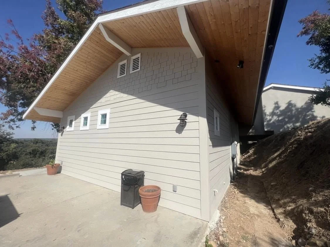 A beige house with a sloped wooden roof, three small windows, and a side wall with a wall-mounted light fixture, situated on a concrete patio with empty plant pots and a trash bin, under a clear blue sky.