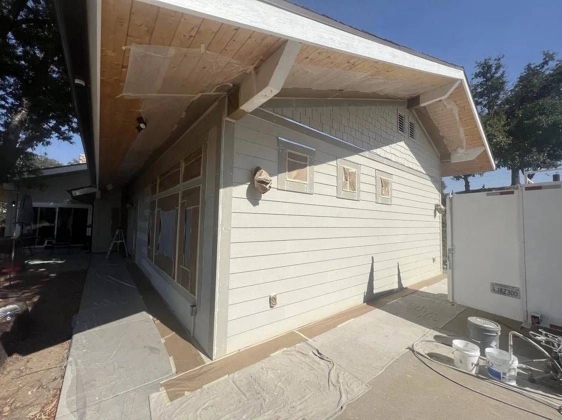 Exterior view of a house under construction with siding and a wooden roof overhang, construction supplies and a trailer nearby.