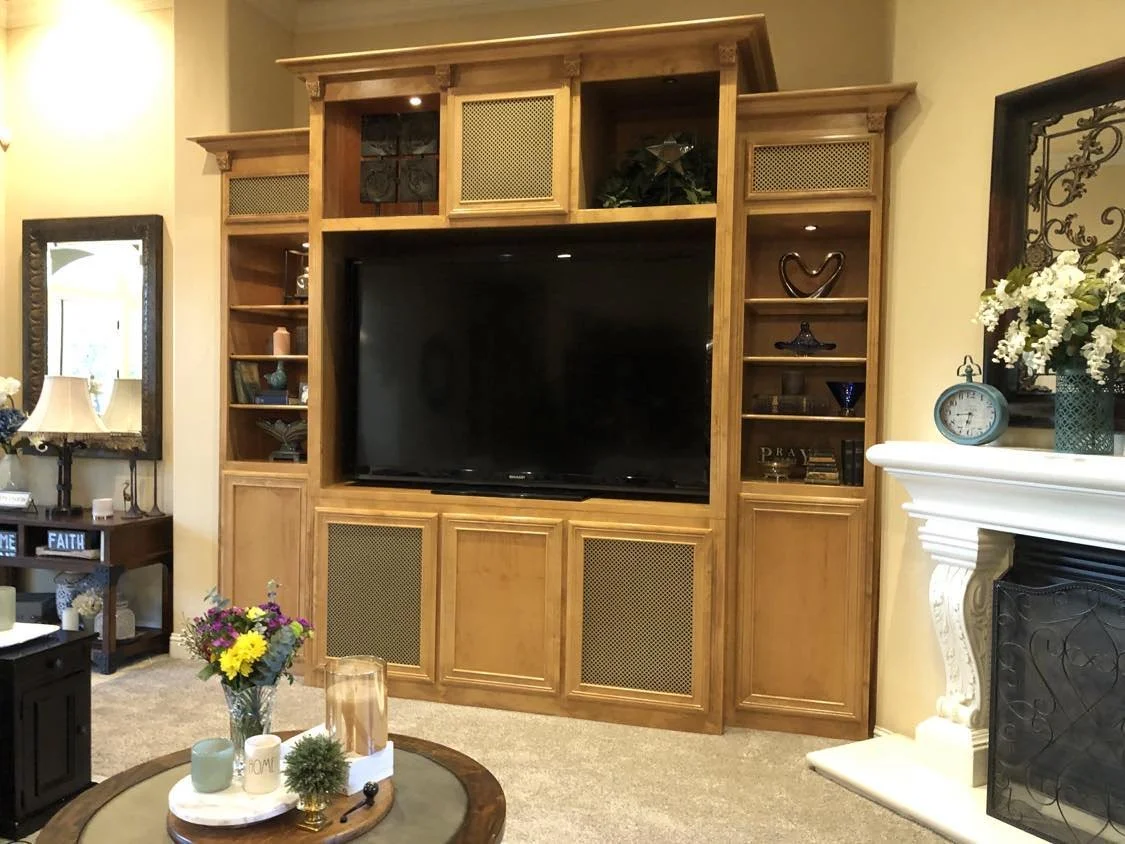 Living room with a large wooden entertainment center housing a flat-screen TV, decorative items, and books, a side table with a lamp and signs reading "Faith" and "Home," a dark coffee table with a floral arrangement, and a white fireplace with a bou