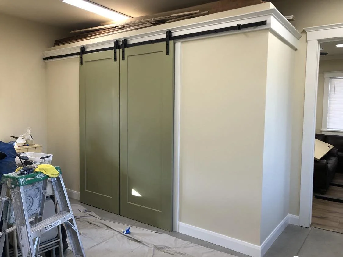 A room with a white-framed sliding barn door on black tracks, partially opened to reveal a beige wall and an adjacent space with black furniture, with some construction tools and materials nearby.