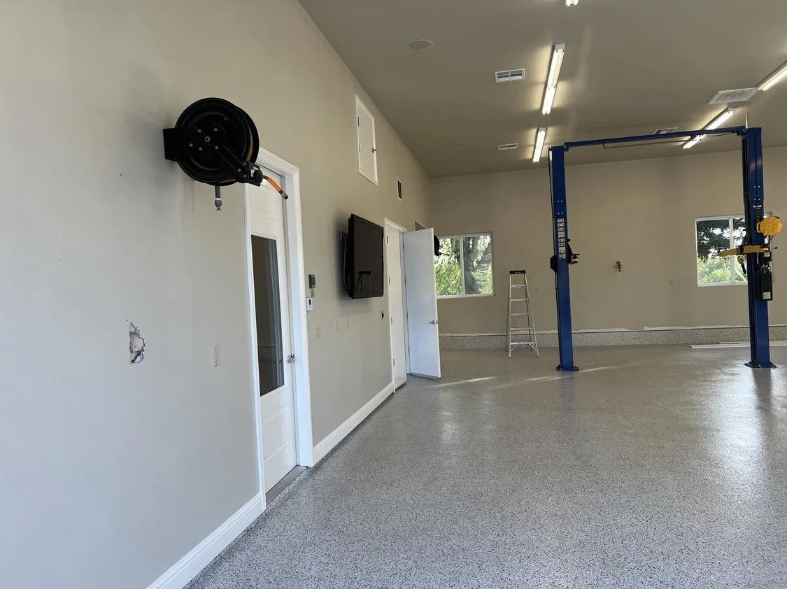 An empty garage or workshop space with a blue vehicle lift, a ladder, a wall-mounted hose reel, and a mounted TV on a beige wall. There are two windows letting in natural light.