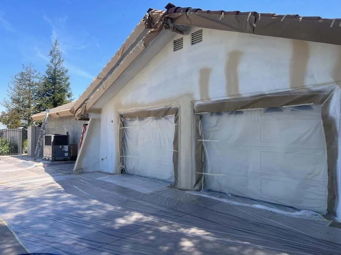 House with garage doors covered in plastic for painting or construction, outdoor area with ladder and construction equipment.
