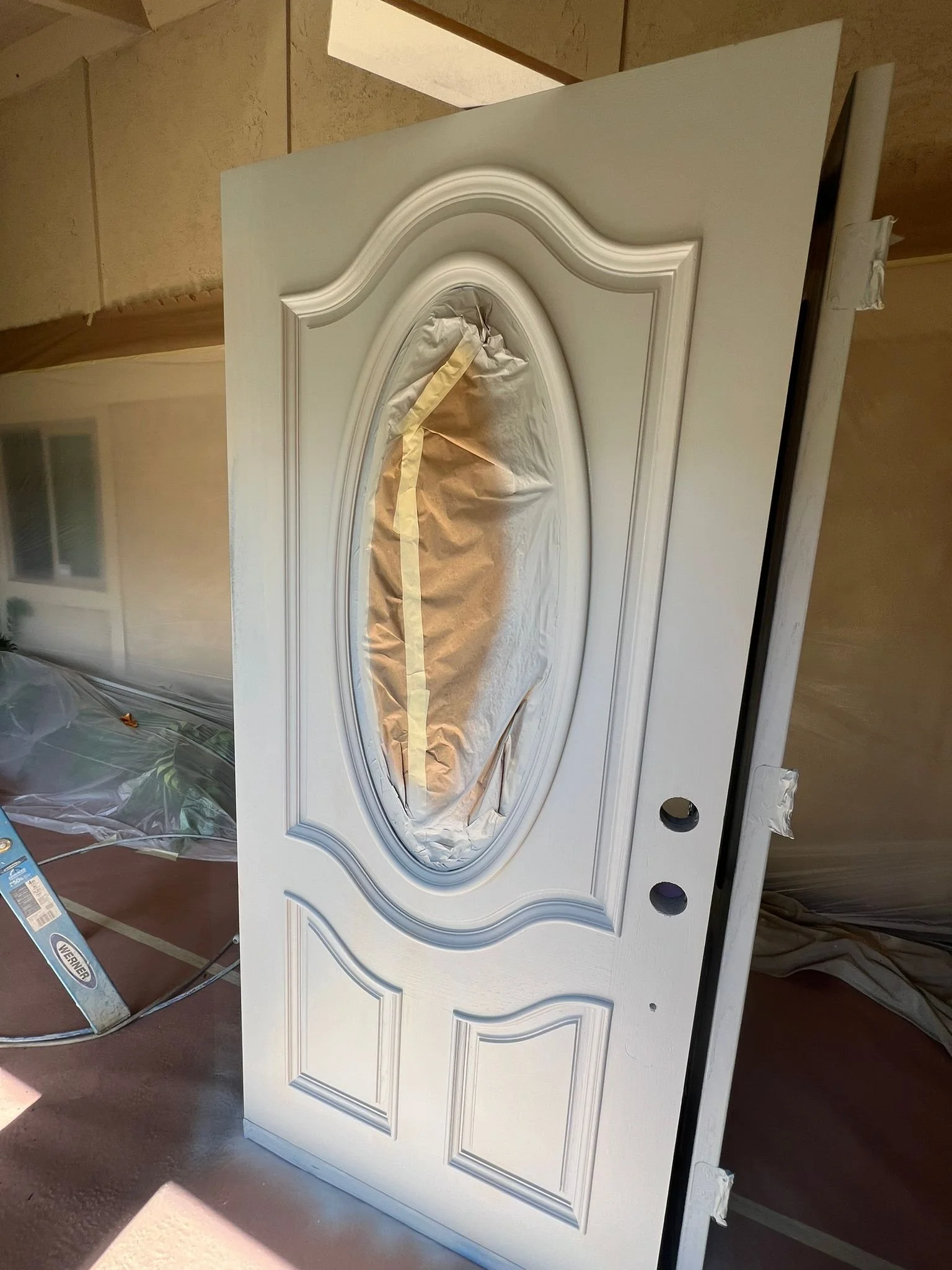 White door with decorative molding, taped off and covered with paper in the oval window area, likely in preparation for painting or finishing.