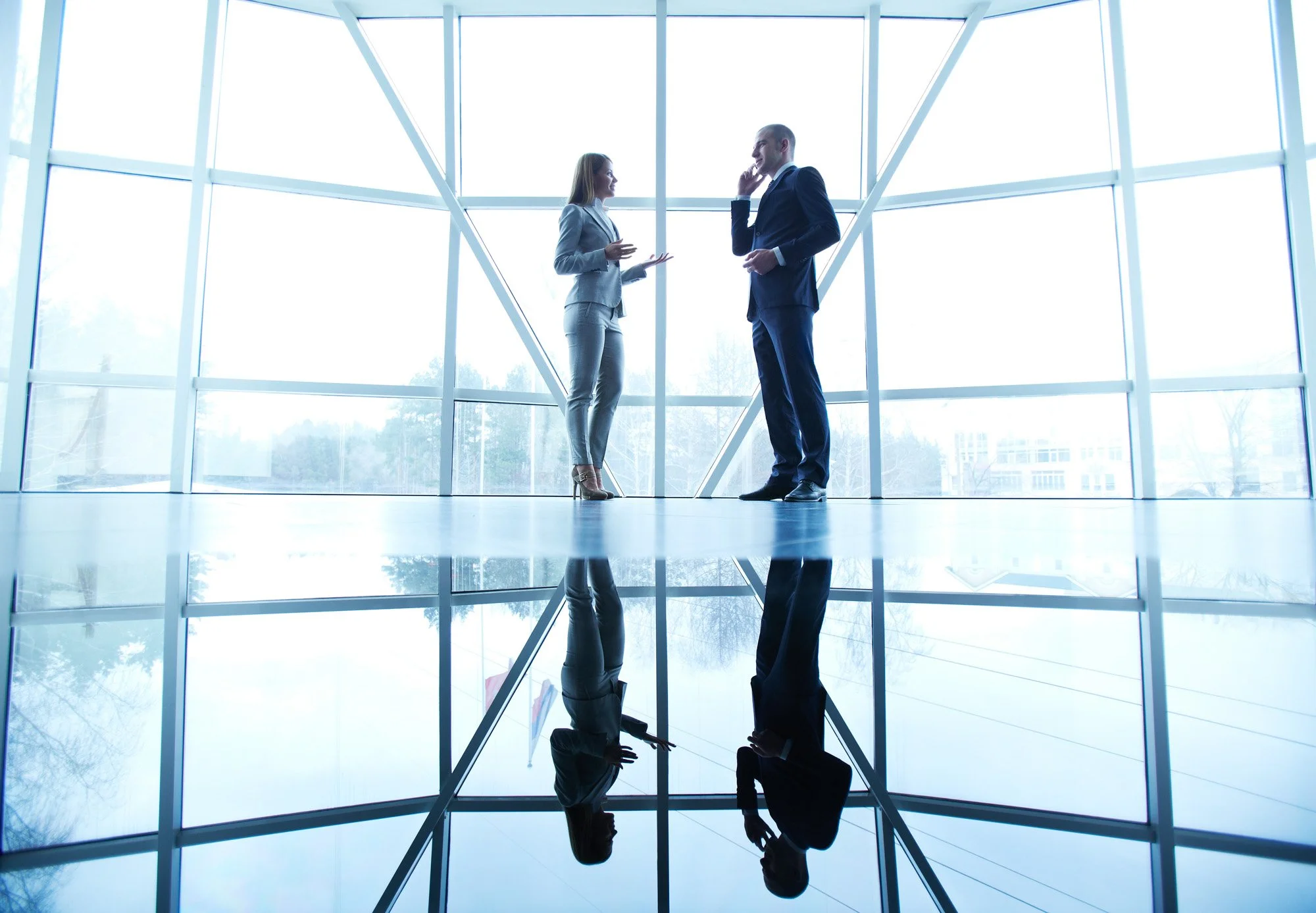 Two business professionals, a woman and a man, having a conversation in a modern office building with large glass windows. Their reflection is visible on the shiny floor.