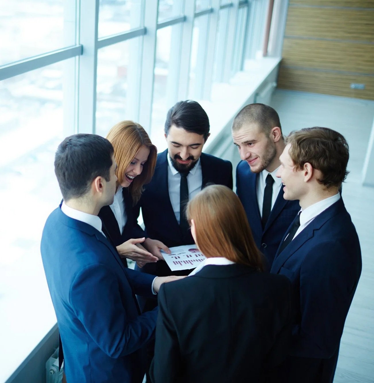 A group of six business professionals, four men and two women, standing in a circle and smiling while looking at a tablet. They are dressed in formal suits and are inside a modern building with large windows.
