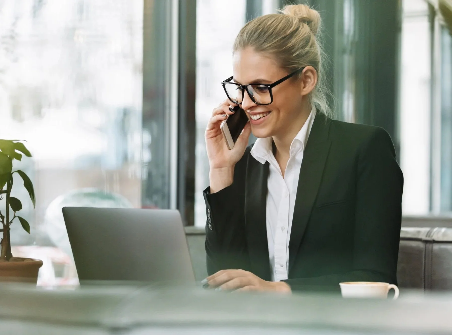 Businesswoman in black blazer and white shirt sitting at a table in a bright office, talking on cellphone, with a laptop, a potted plant, and a cup on the table.