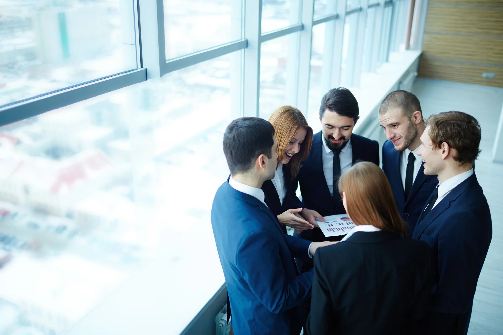 Group of six business professionals in suits standing together, looking at a tablet and smiling, in a modern office with large windows.