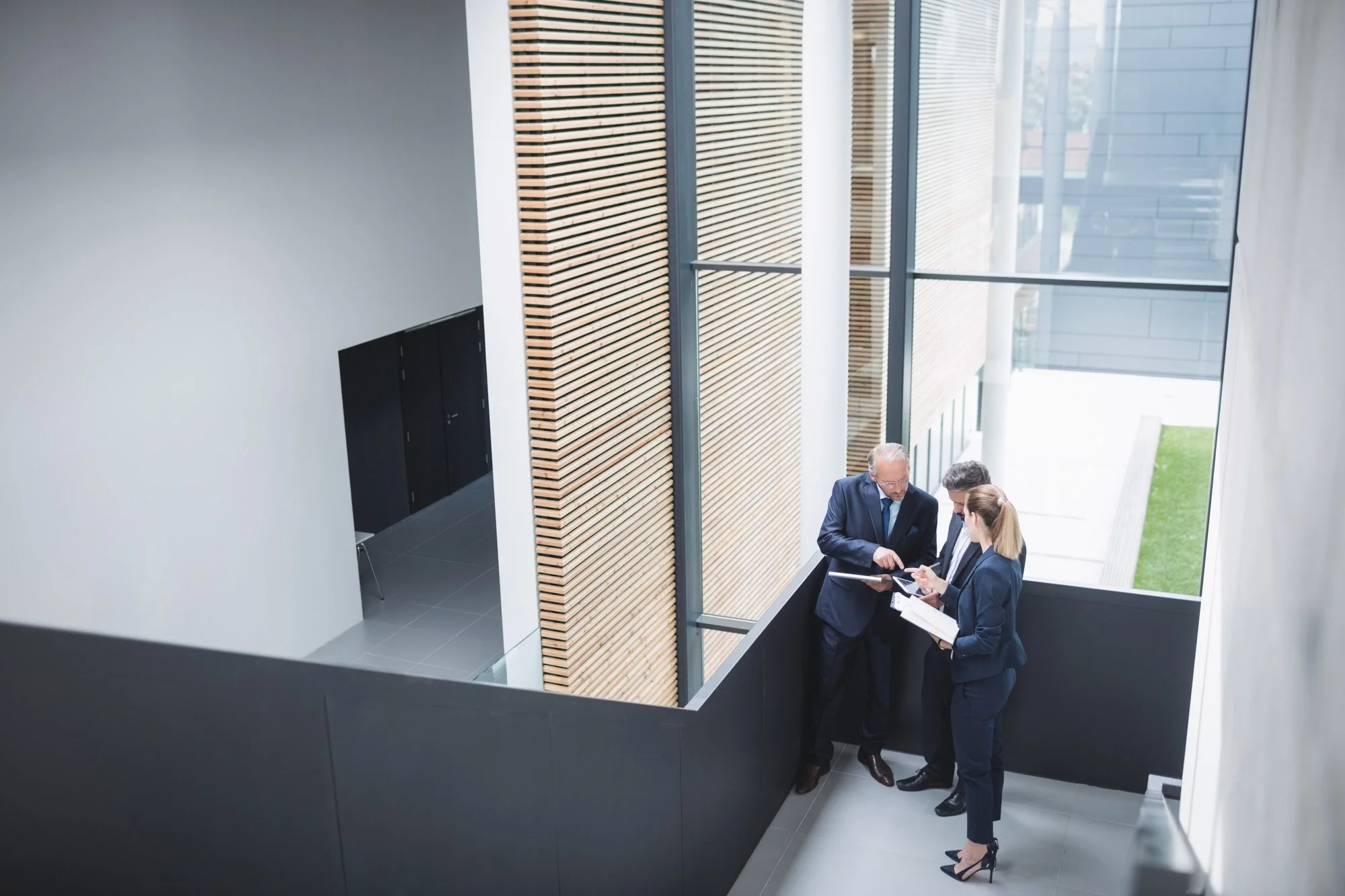 Three business professionals, two men and one woman, are standing on a balcony inside a modern office building, engaged in a discussion and looking at documents.