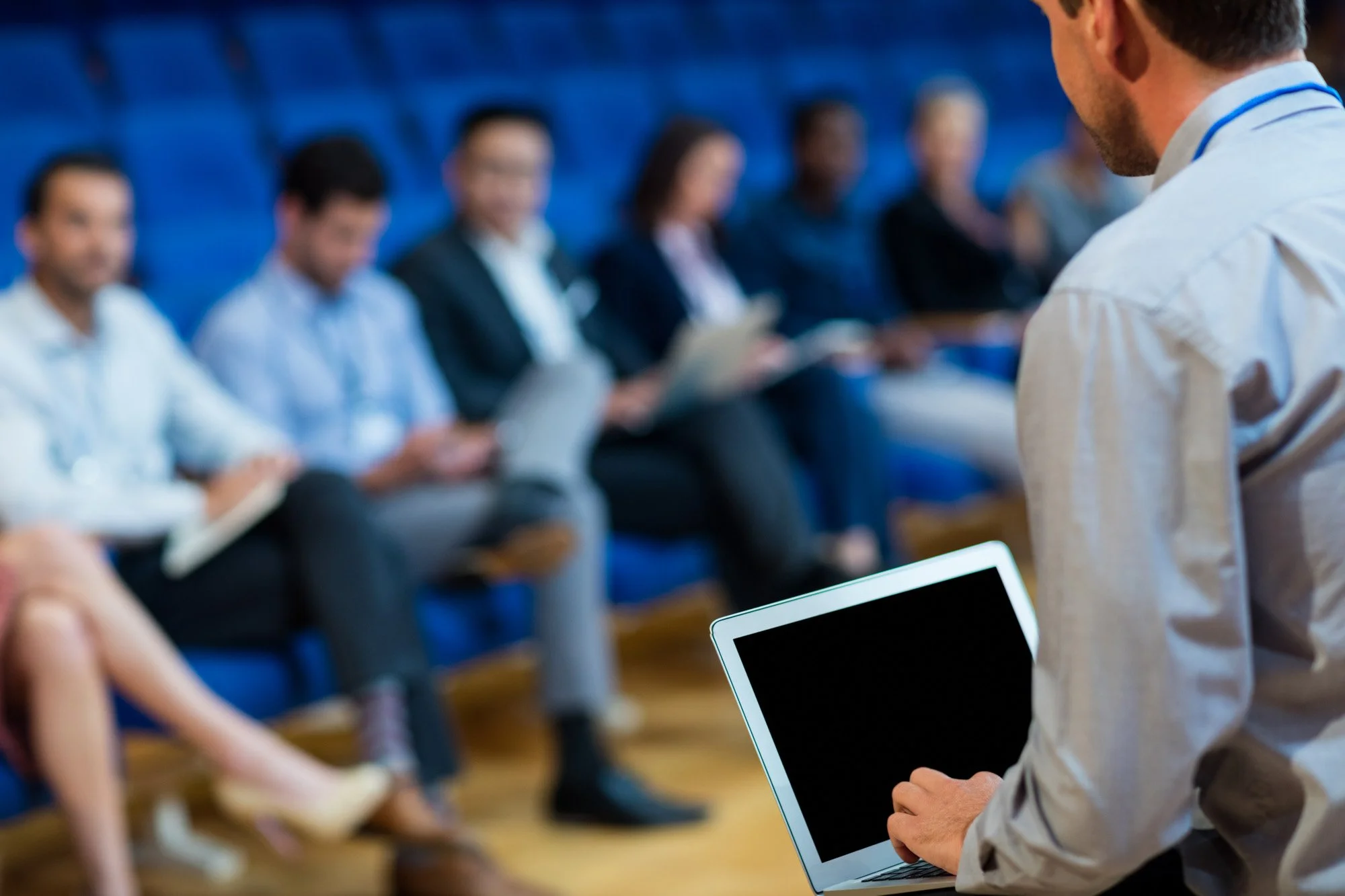 A person giving a presentation to a group of diverse professionals seated in an auditorium, holding a tablet and facing a screen.
