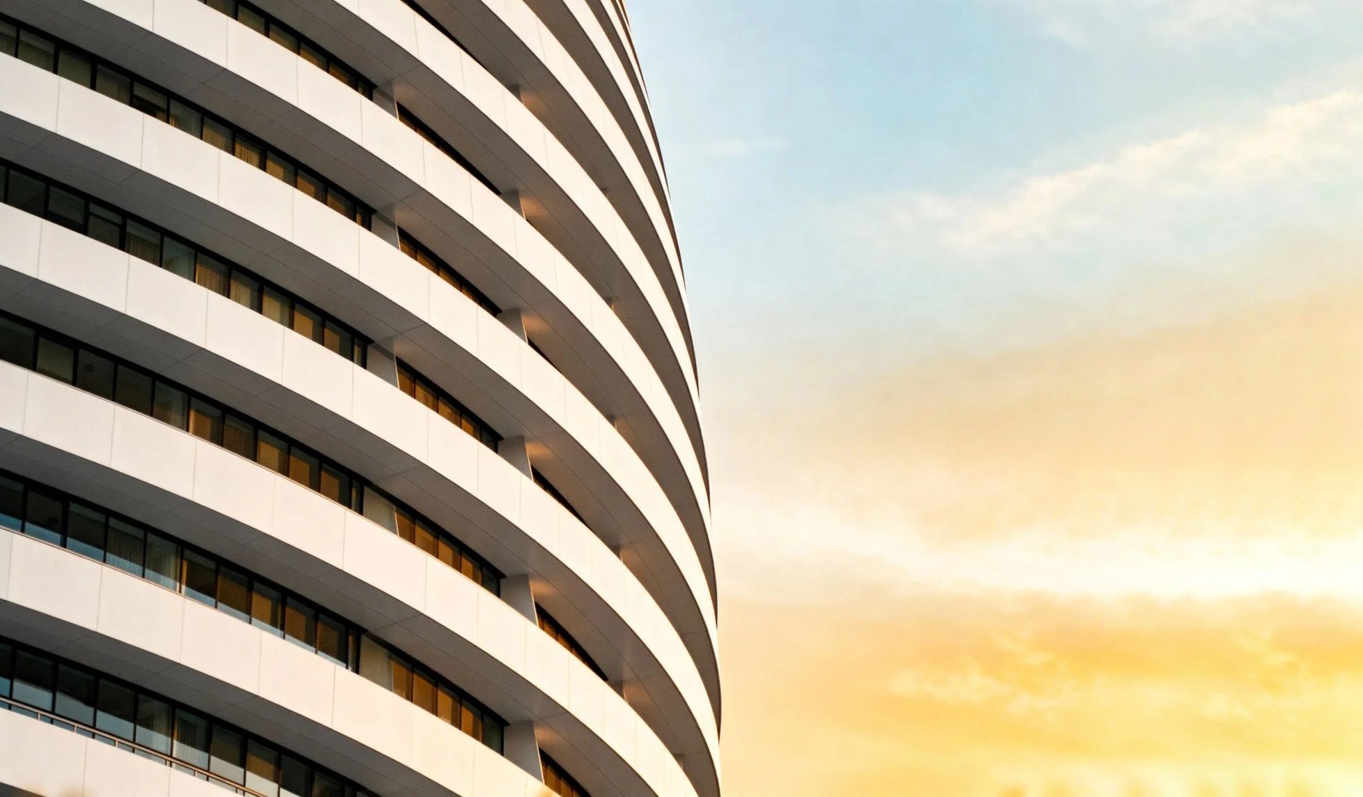 Close-up view of a modern, curved high-rise apartment building with white balcony railings and glass windows at sunset.