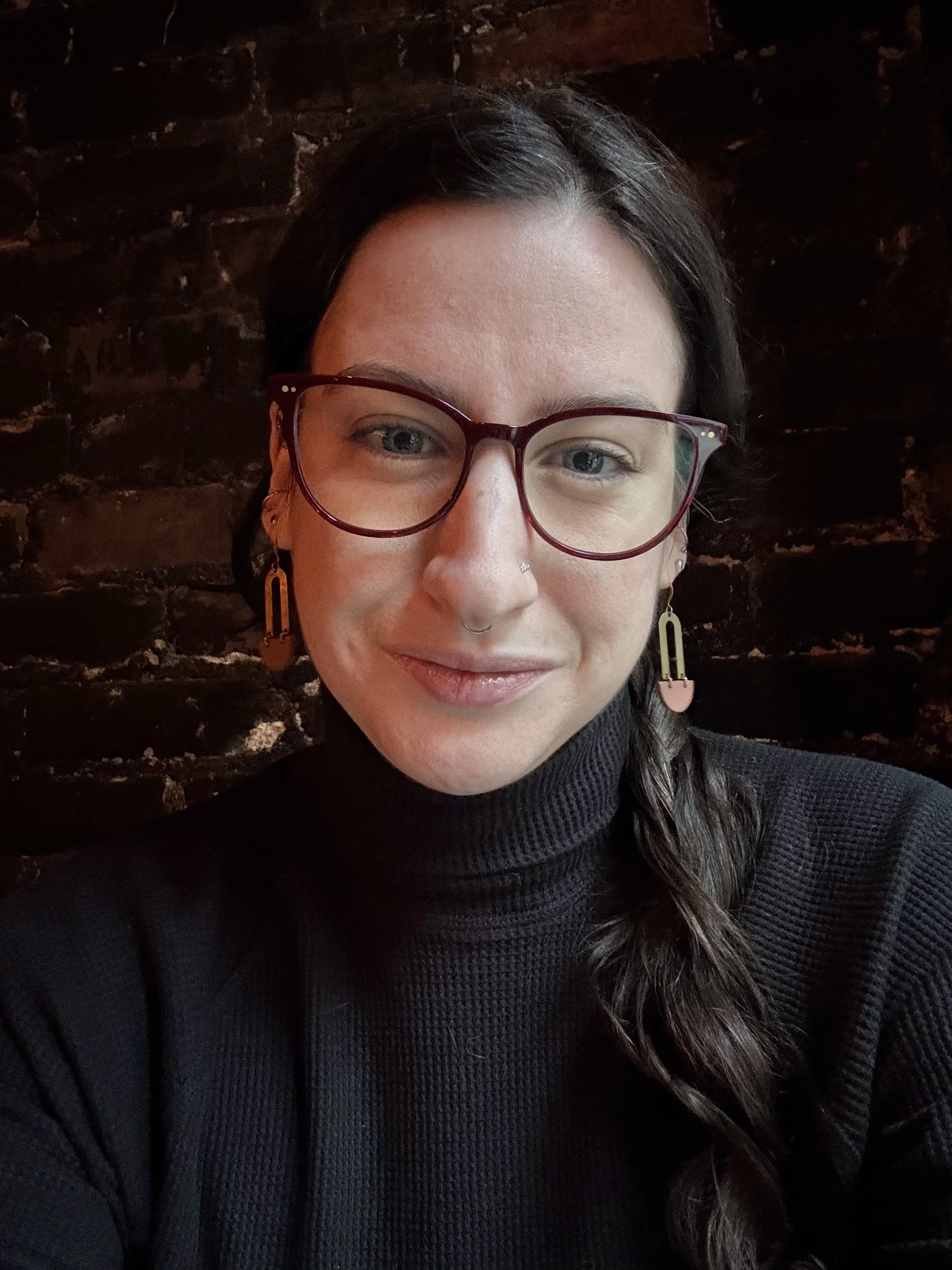 A woman with long dark hair, wearing glasses, earrings, and a black turtleneck, smiling and looking at the camera in front of a dark brick wall background.