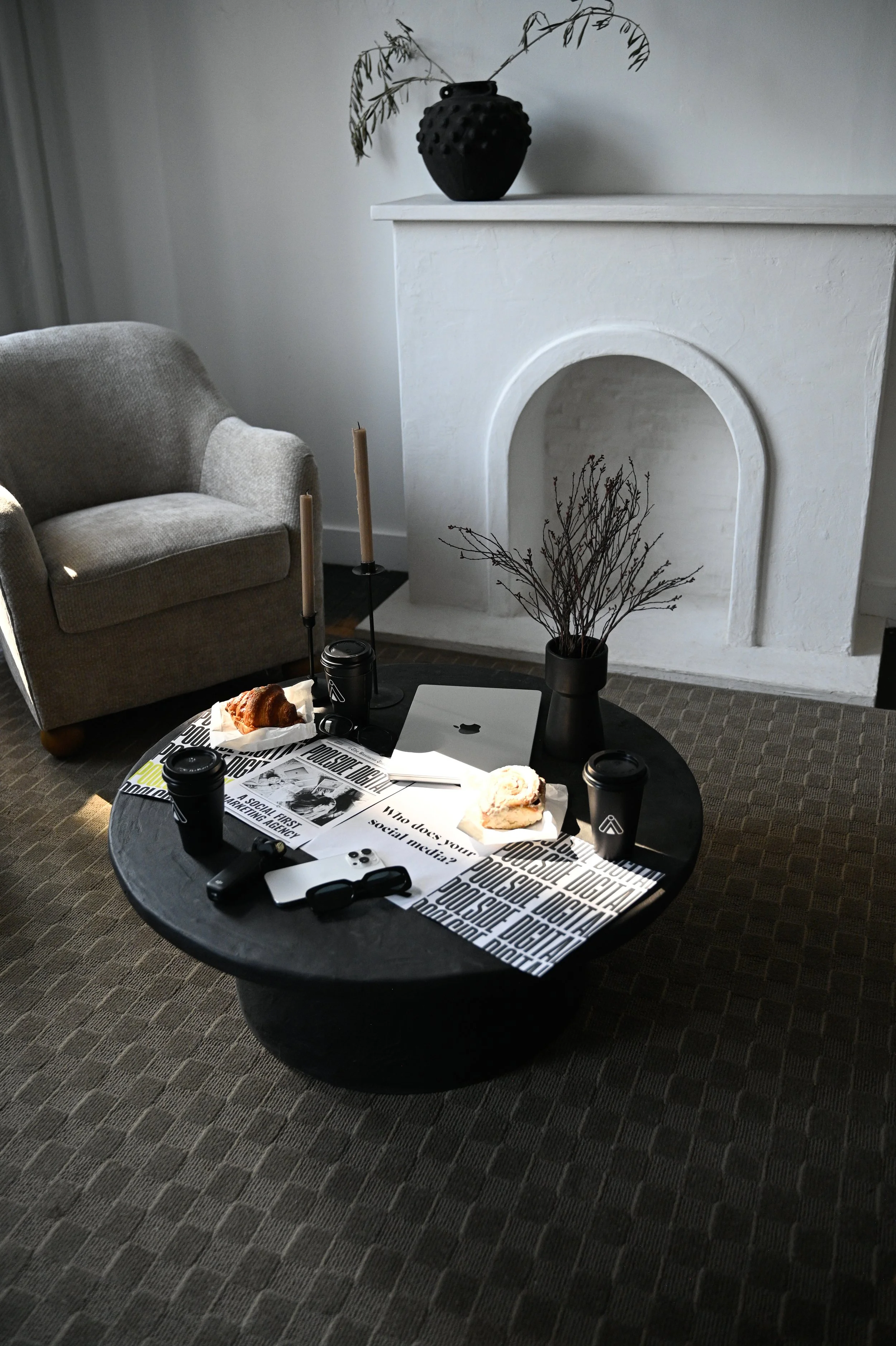 Living room with a black coffee table, beige armchair, white fireplace, and decorative items including a black vase with branches, candles, and food on paper plates, with coffee cups and glasses on the table.