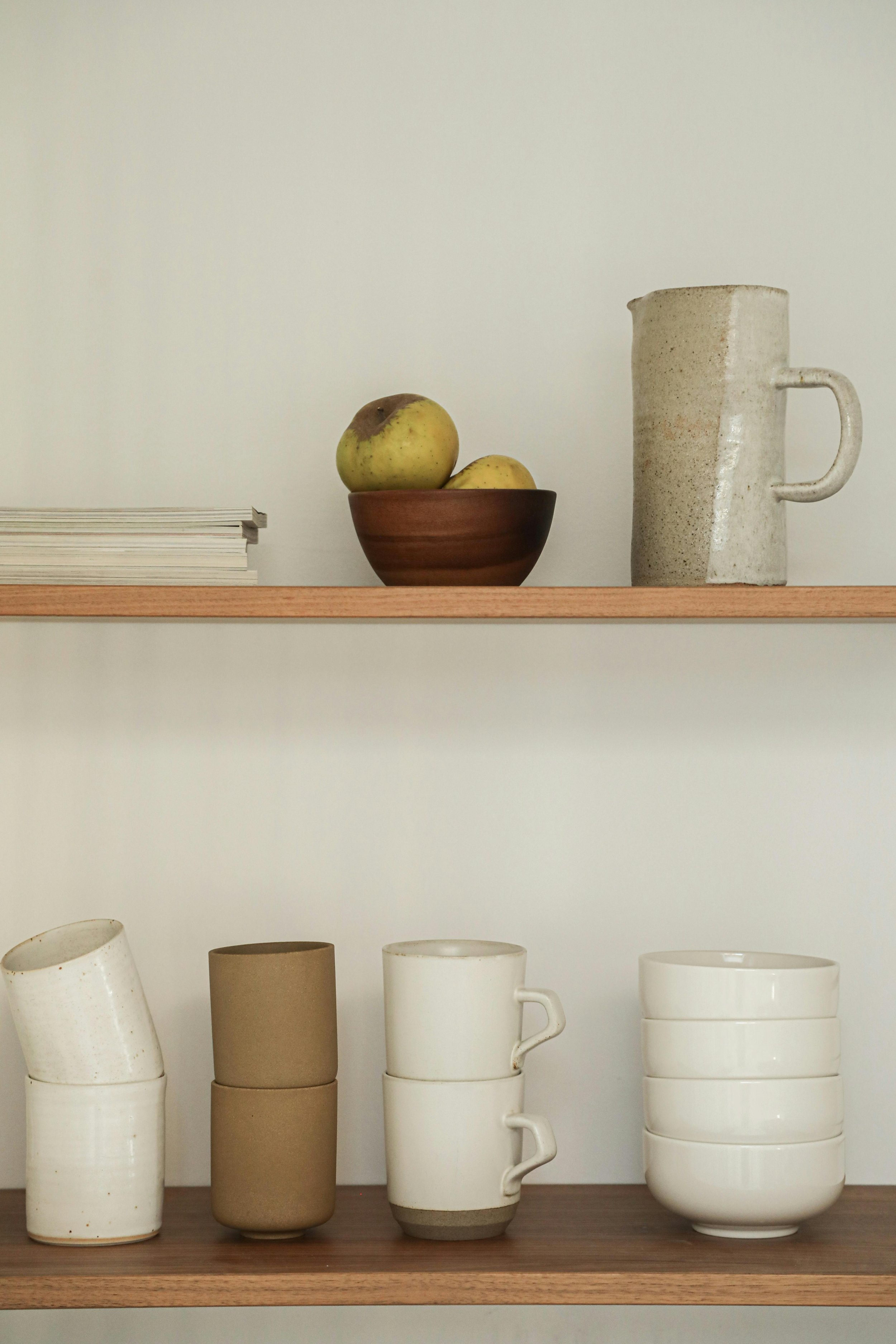 A minimalist wooden shelf displaying ceramic dishes, bowls, and a coffee mug, with a plain wall background.