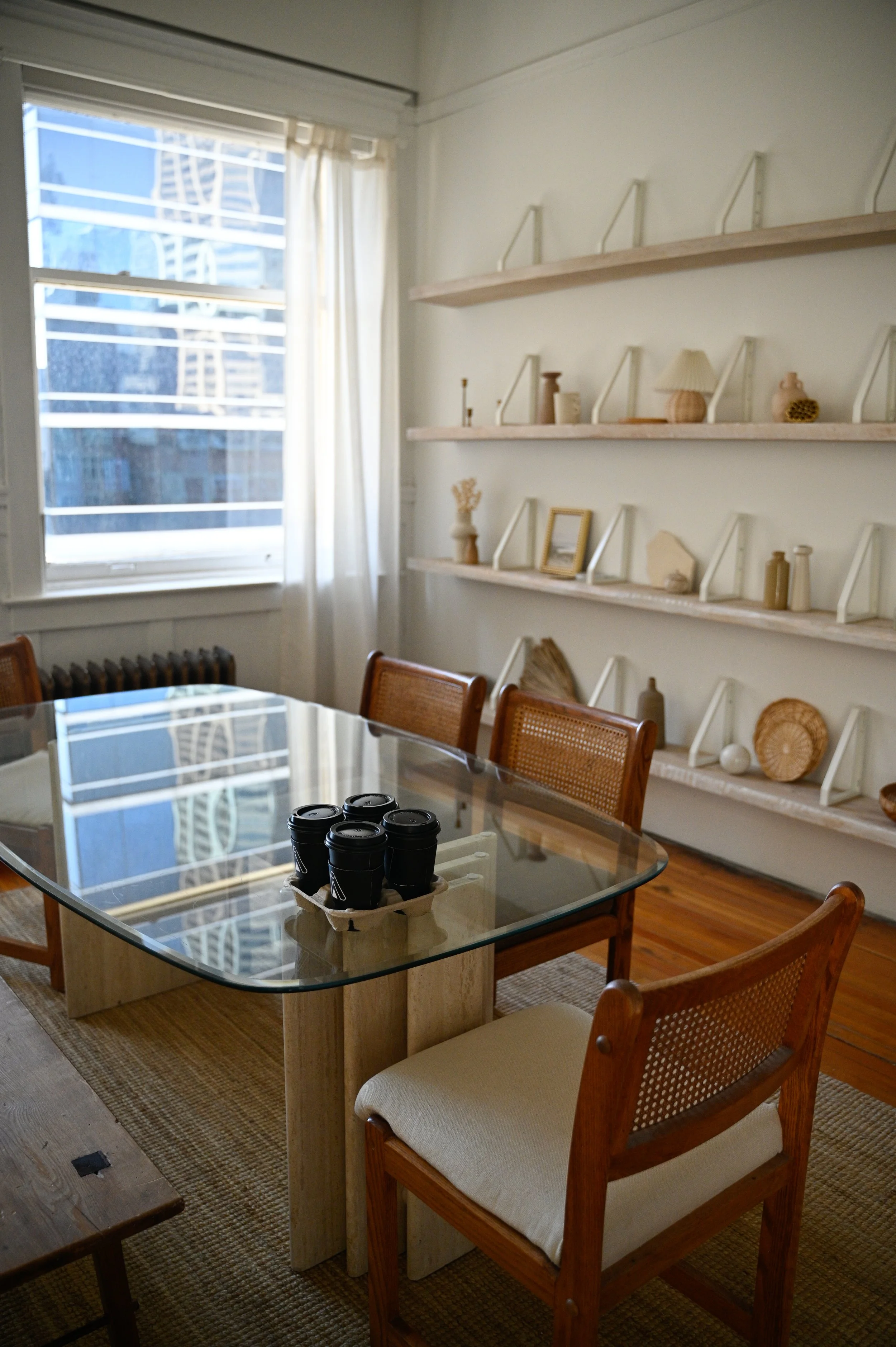 A cozy dining room with a glass-top table, wooden chairs, and white shelves on the wall decorated with vases, framed pictures, and decorative objects, next to a window with white curtains.