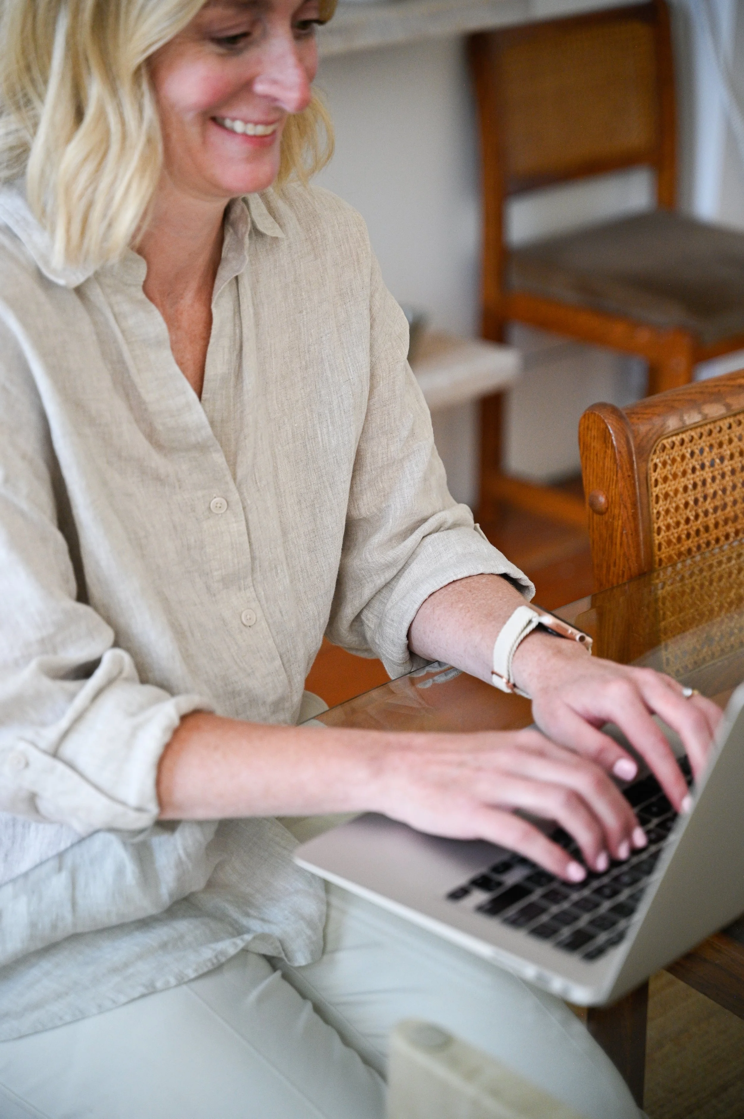 A woman with blonde hair wearing a beige button-up shirt working on a laptop at a wooden table.