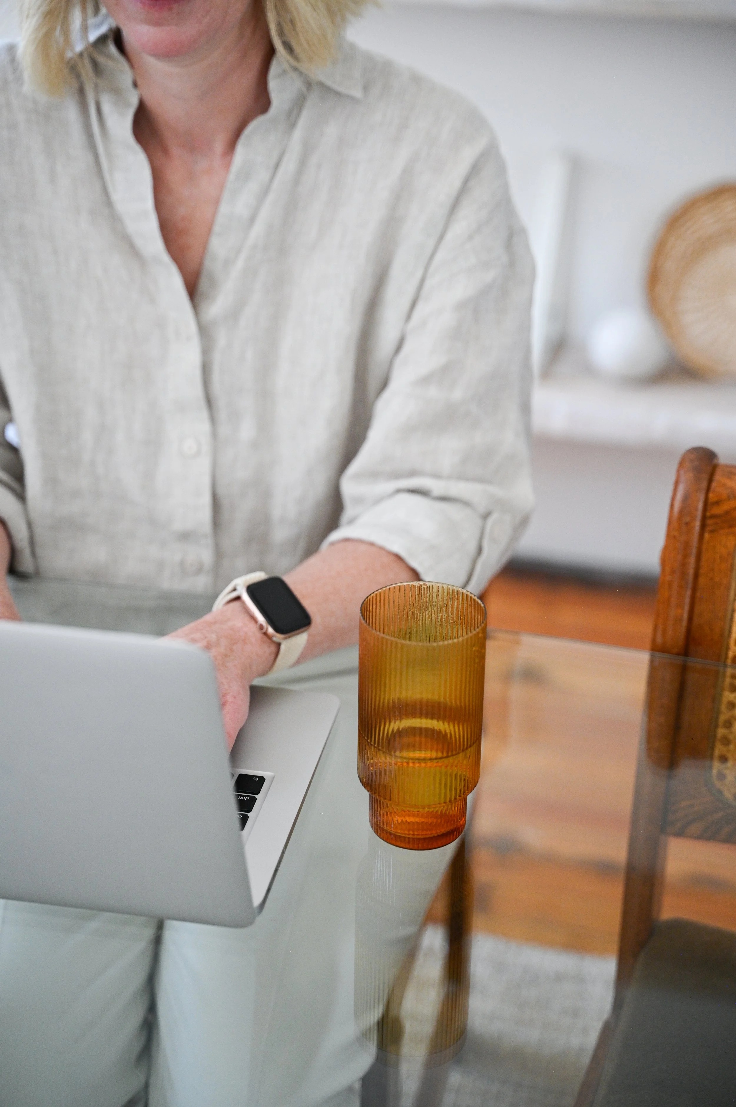 A woman sitting at a dining table with a laptop, wearing a beige shirt and a smartwatch, with a large amber-colored glass on the table.