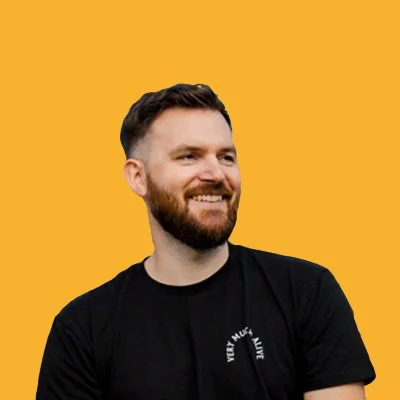 A smiling man with a beard and short hair, wearing a black T-shirt, against a yellow background.