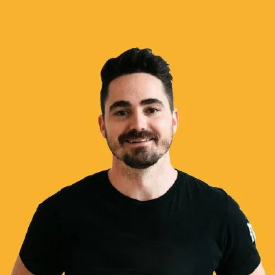 A man with dark hair and a beard, smiling, wearing a black t-shirt, standing against a yellow background.