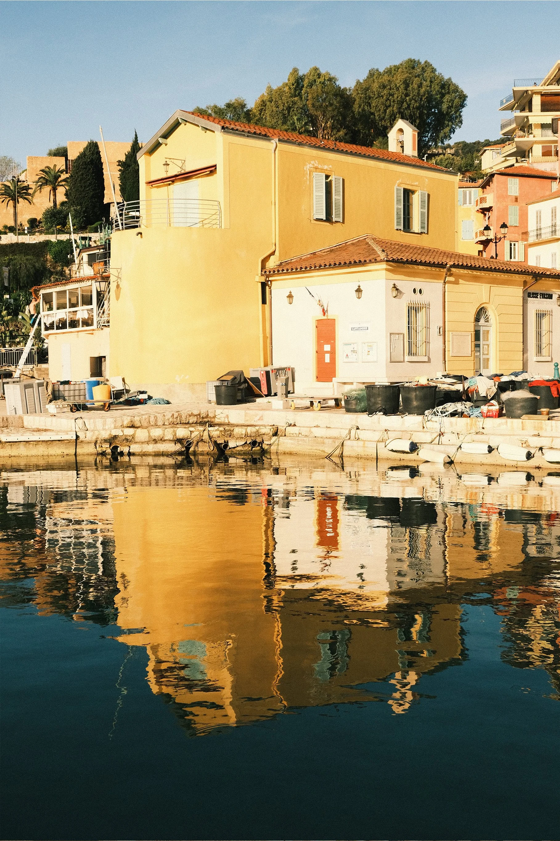 Maisons colorées au bord de l'eau avec leur reflet dans la rivière, ensoleillées, avec des collines en arrière-plan.
