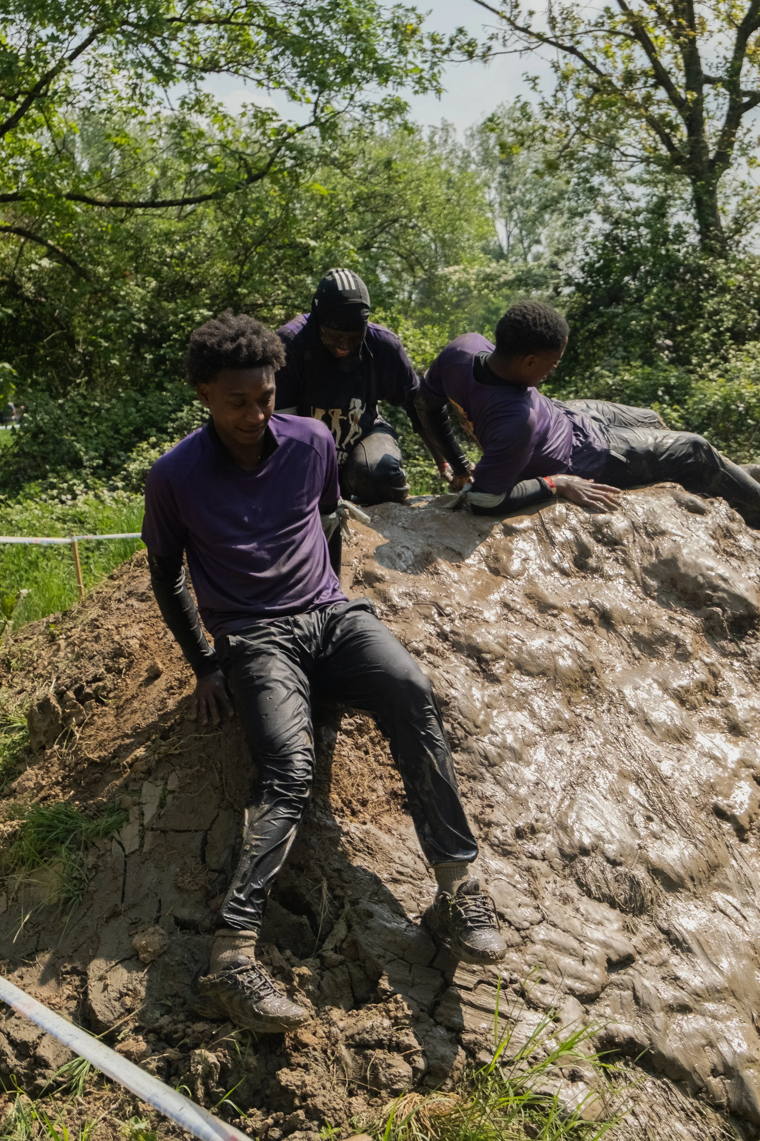 Trois jeunes hommes en vêtements de sport sont en train de grimper ou de descendre une surface boueuse dans une zone boisée, lors d'une activité de sport ou d'aventure.