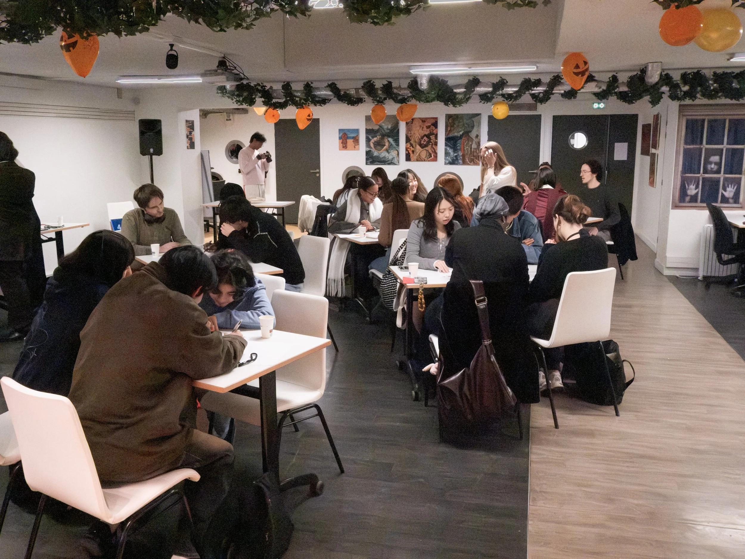 Groupe de personnes assises à des tables dans une salle décorée avec des ballons d'Halloween orange et des guirlandes vertes, participants à une activité ou une réunion, avec quelques personnes en train de prendre des photos ou de parler.