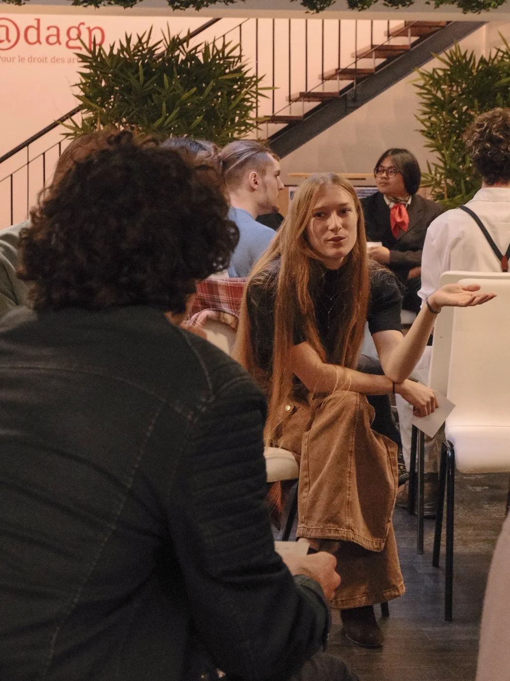 Groupe de personnes en discussion dans une salle avec plantes vertes et escaliers en arrière-plan.