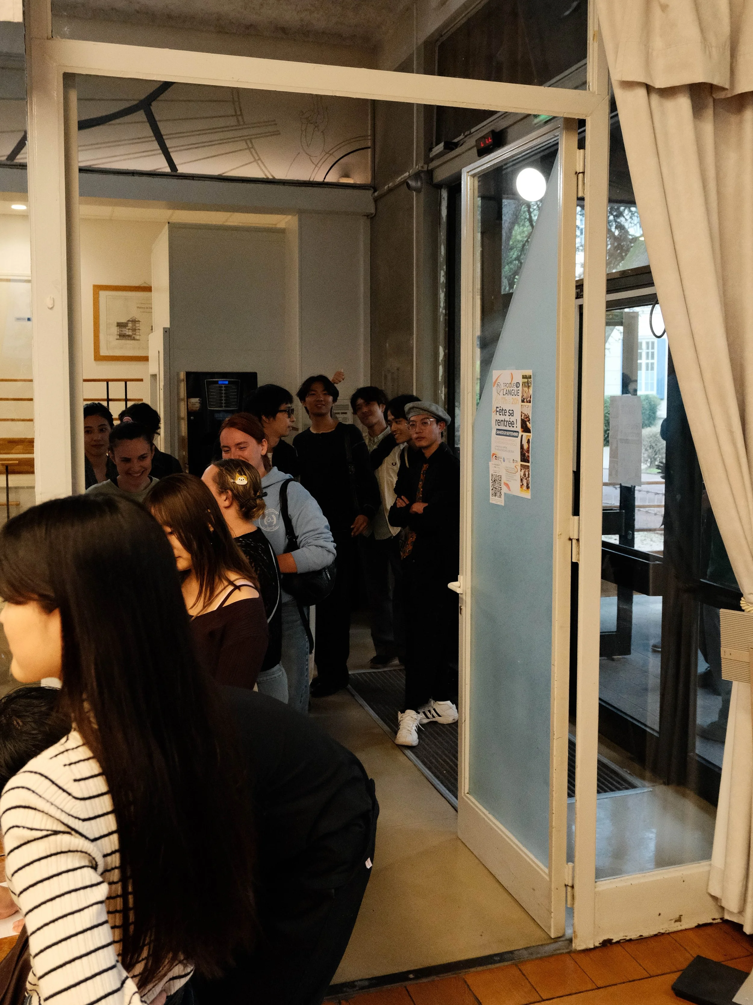 Groupe de jeunes attendant à l'entrée d'un bâtiment, certains discutant et souriant, à l'intérieur d'un lieu accueillant.