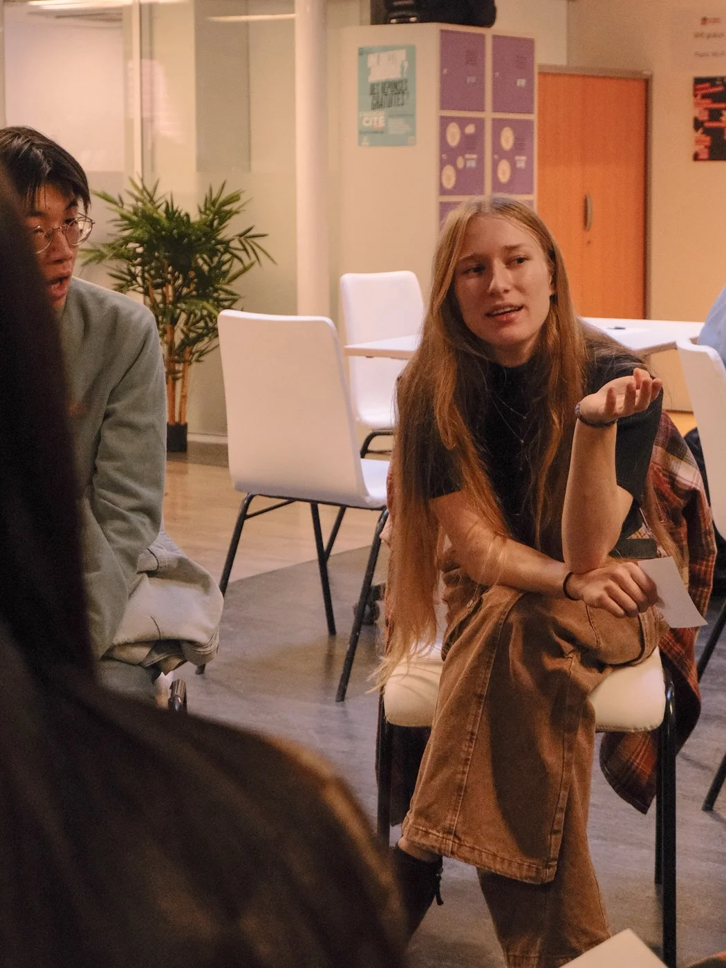 Une jeune femme rousse parle lors d'une discussion ou d'un atelier, assise sur une chaise dans une salle de réunion ou de classe, entourée d'autres personnes.