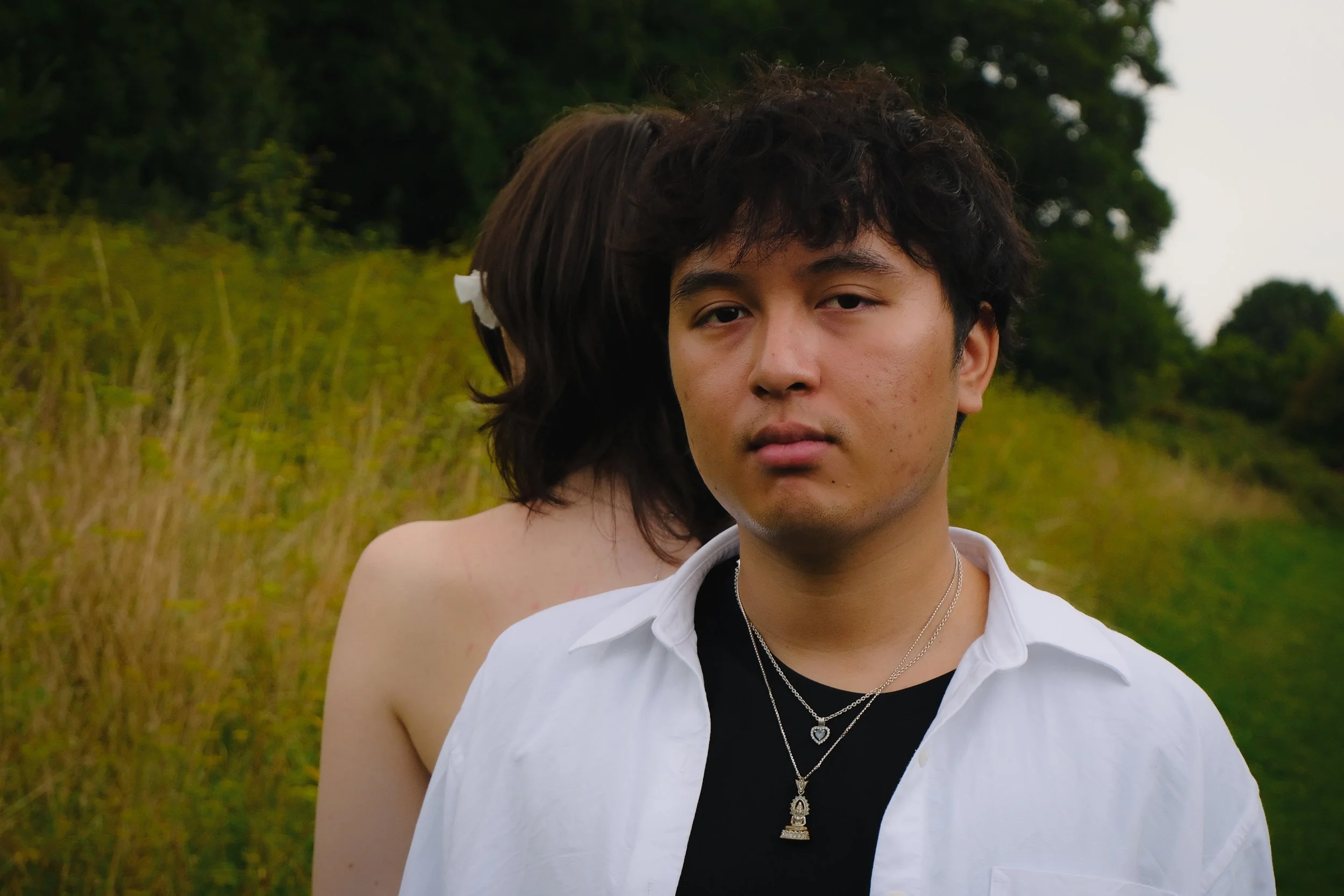 Un jeune homme debout devant un paysage naturel, avec une femme derrière lui dont le visage n'est pas visible.