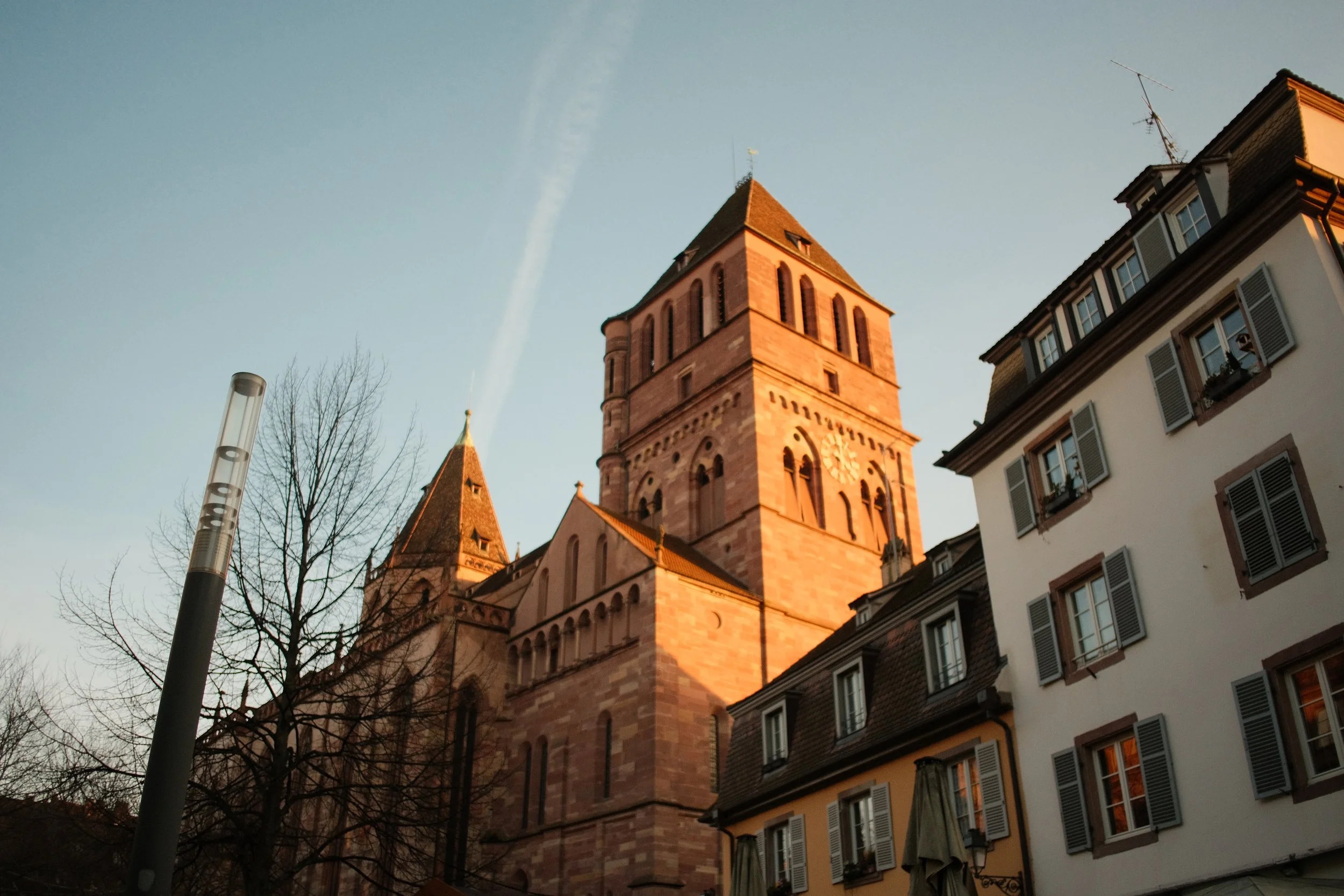 Une photo d'une vieille église en pierre, avec un clocher, prise au coucher du soleil. Des bâtiments avec des fenêtres à volets autour, un arbre sans feuilles et un lampadaire sont aussi visibles.