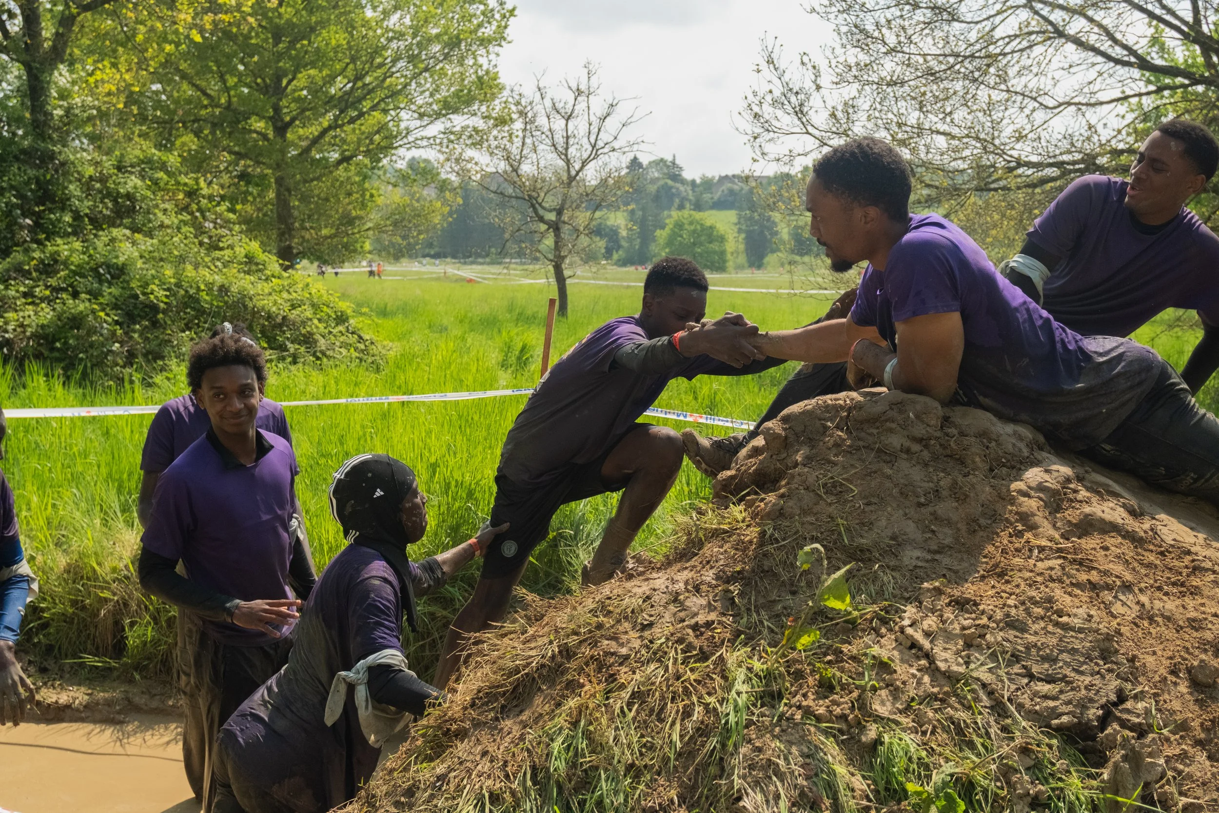 Groupe de jeunes aidant à sortir un participant d'une zone boueuse lors d'une course ou un défi en plein air dans un parc vert avec des arbres et une étendue d'herbe.